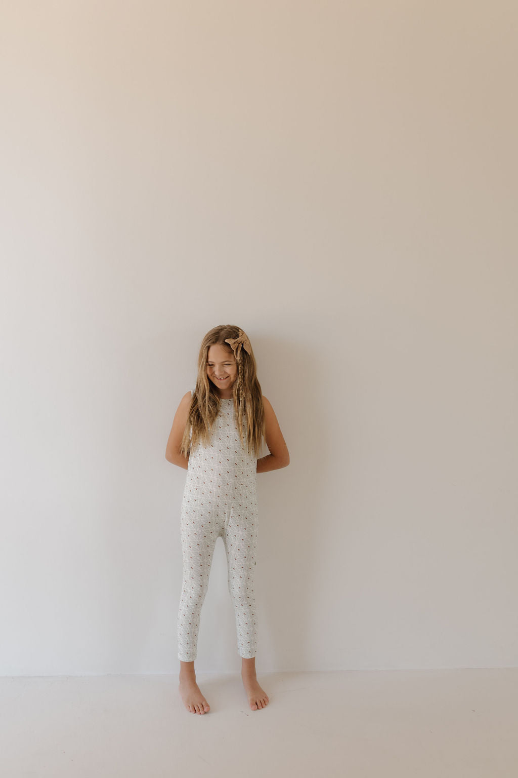 A young girl with long hair stands barefoot against a plain wall, wearing the Harem Bamboo Romper | Coast by forever french baby. The white, sleeveless romper features a subtle pattern and breathable fabric for comfort. She has a bow in her hair and her hands behind her back.
