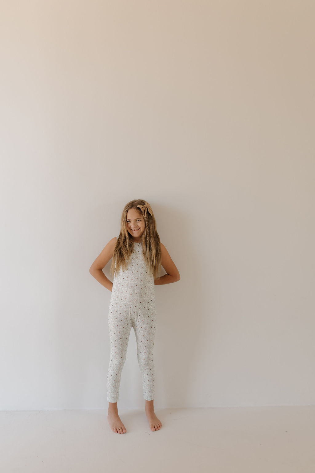A young child with long hair stands barefoot against a plain, light-colored wall, wearing the sleeveless Harem Bamboo Romper in Coast by forever french baby. The matching headband and minimalistic setting highlight the serene vibe of the breathable, hypo-allergenic clothing.