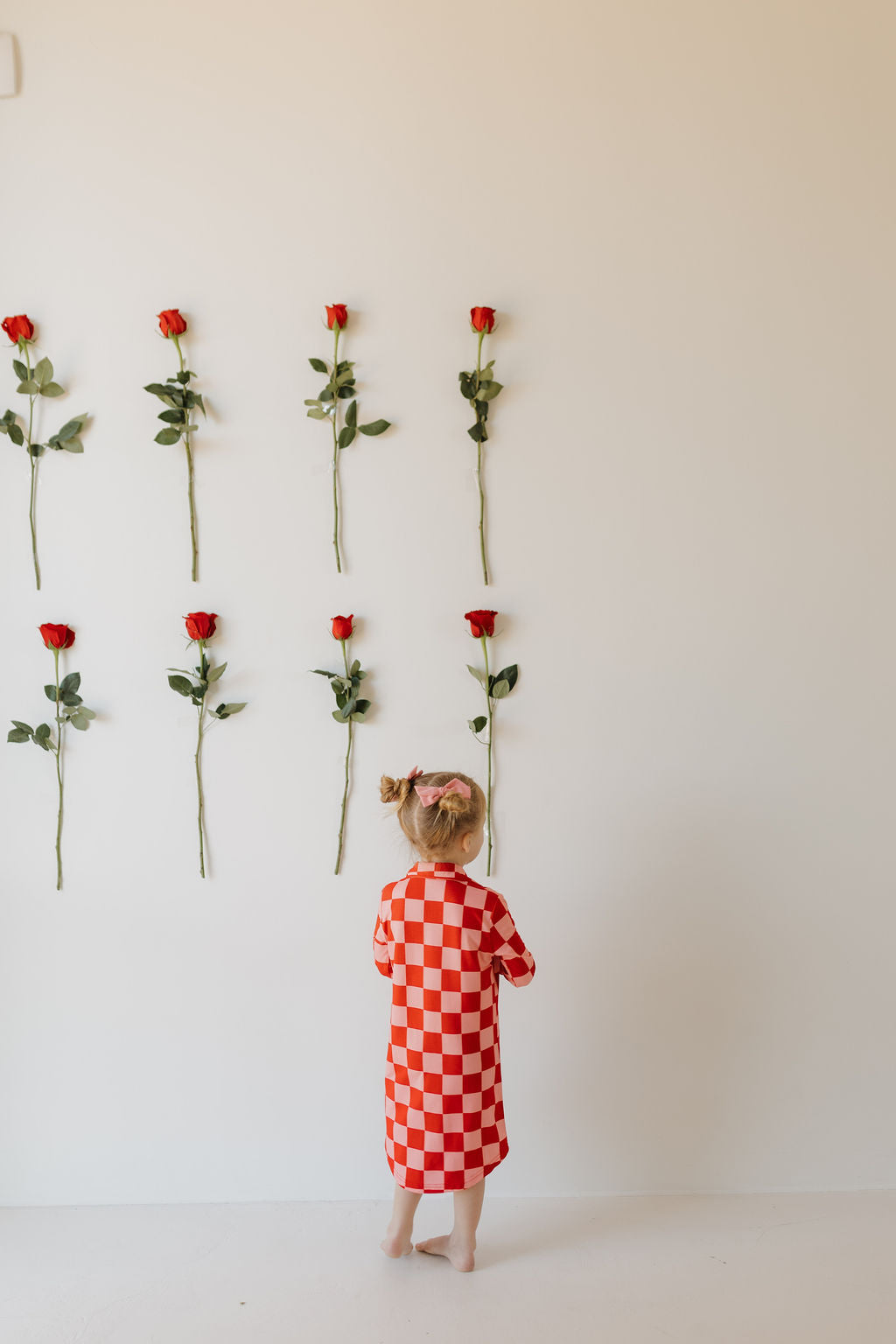 A child with blonde hair stands barefoot, wearing the Toddler Bamboo Sleeping Dress | XOXO by forever french baby, made from hypo-allergenic material. They face a white wall decorated with six red roses arranged in vertical rows.