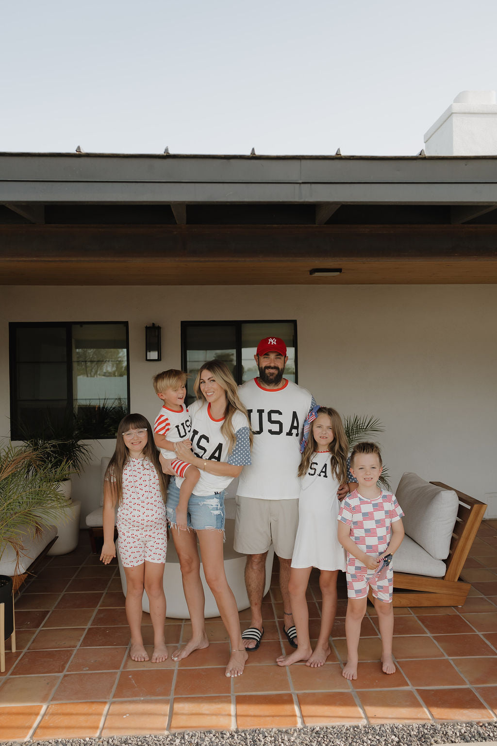 A family of five smiles on a patio, wearing forever french baby Adult Bamboo T-Shirts | Stars & Stripes—casual and breathable summer outfits with “USA” and patriotic patterns—gathered by an outdoor seating area.