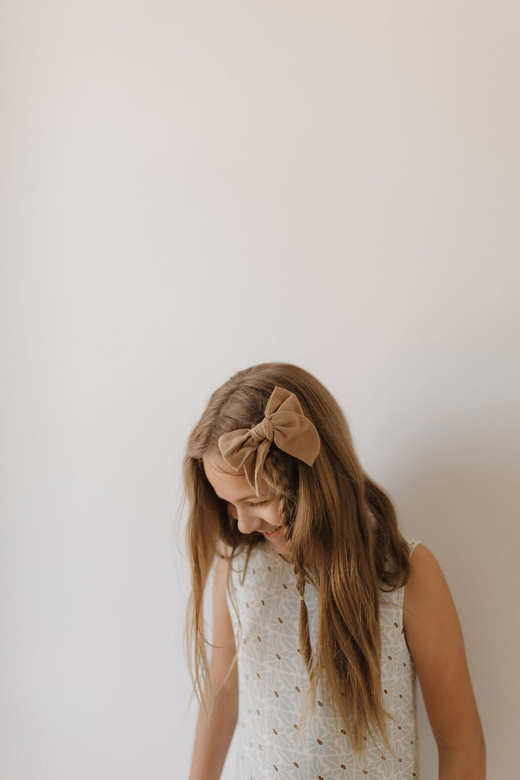 A young girl with long brown hair stands against a plain white background, wearing the Harem Bamboo Romper by forever french baby, crafted from breathable fabric and adorned with delicate patterns. She has a large tan bow in her hair.