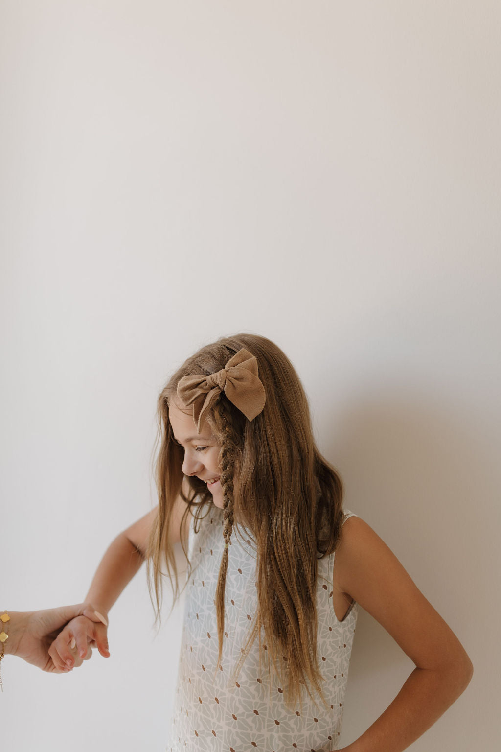 A young girl in a sleeveless Harem Bamboo Romper by forever french baby is smiling and looking down, her long braided hair adorned with a large bow headband. Her left hand is gently held by an unseen person against a plain white background.