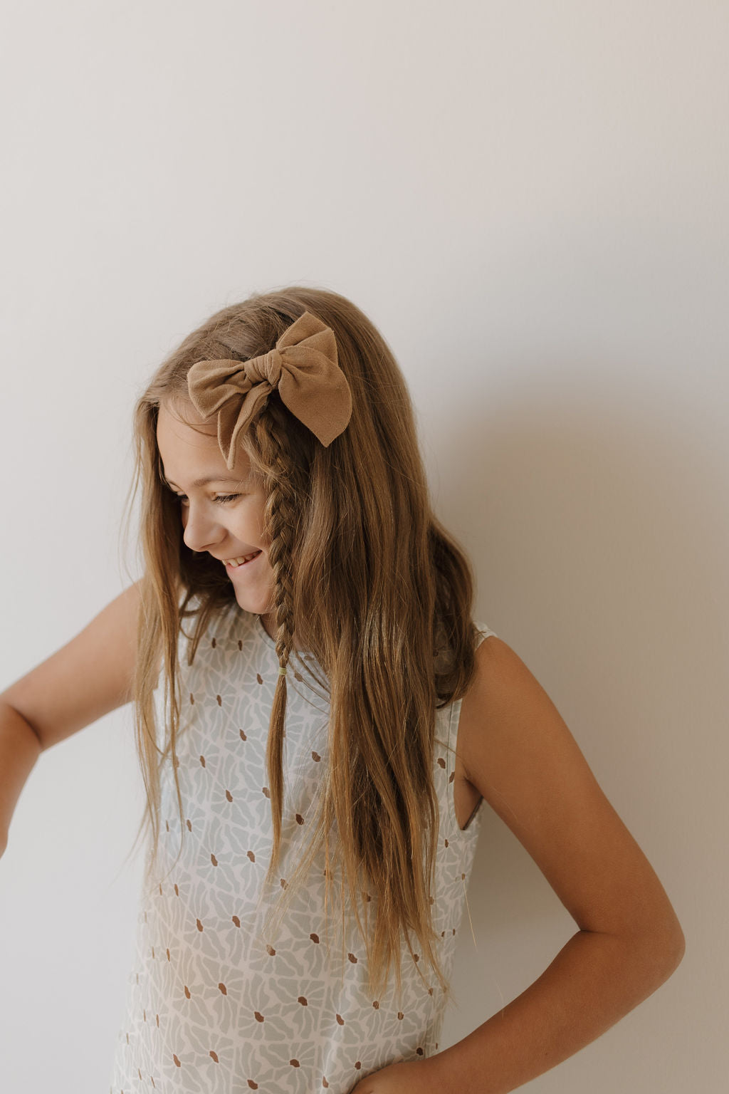 A young girl with long brown hair is wearing a sleeveless patterned Harem Bamboo Romper | Coast by forever french baby. She has a large brown bow in her braided hair, smiling as she looks downwards against a light background.