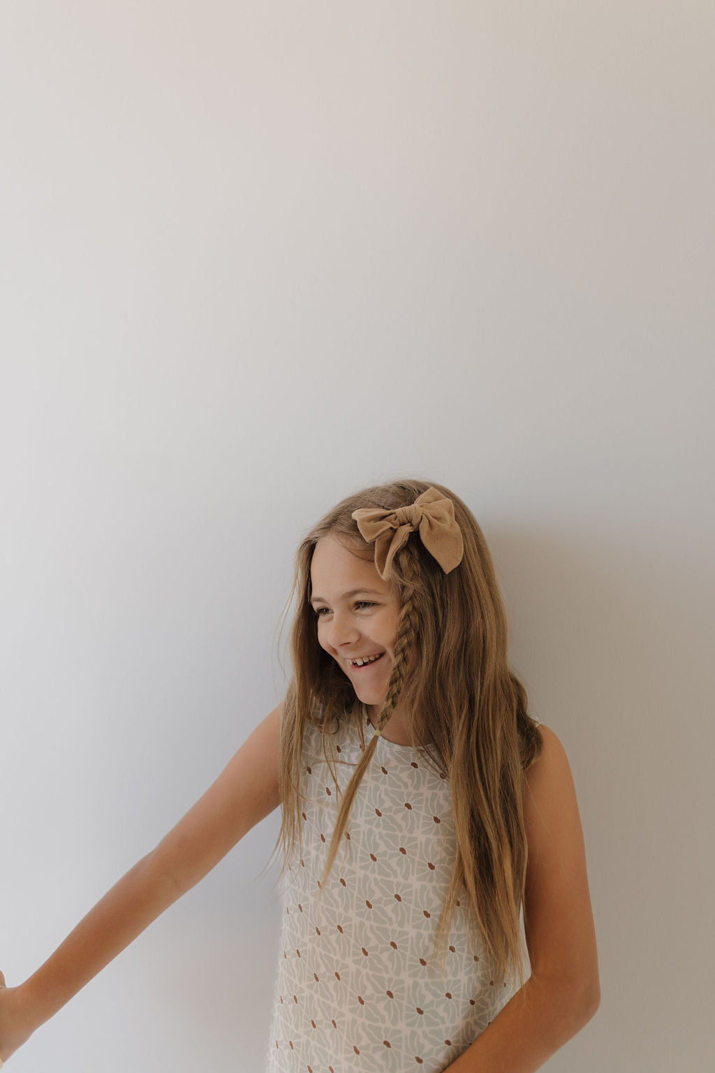 A young girl with long hair, wearing a sleeveless Harem Bamboo Romper by Forever French Baby in breathable fabric, has a brown bow in her hair. She smiles and looks to the side against a plain white background.