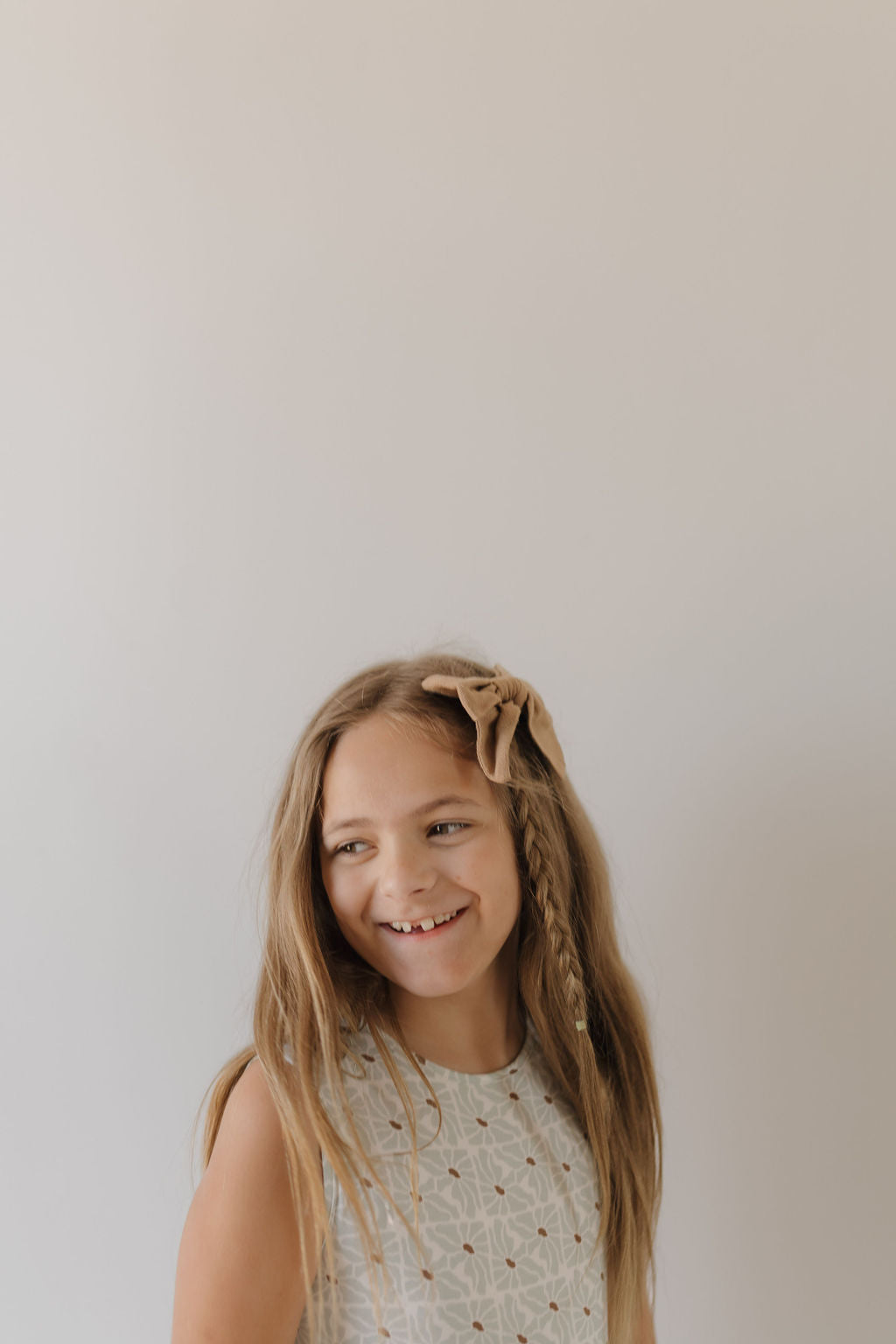 A young girl with long blond hair smiles while looking to the side, her braid decorated with a large bow. She wears the Harem Bamboo Romper from forever french baby, made of breathable fabric. The plain, neutral background highlights her radiant expression.