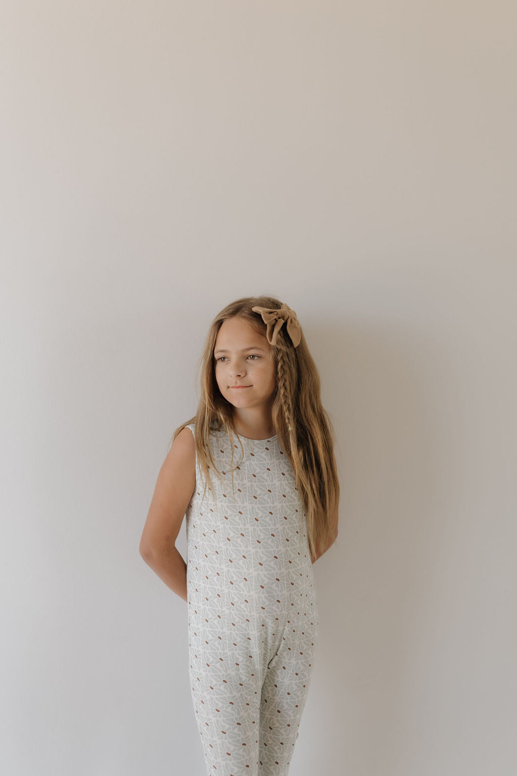 A young girl with long hair and a bow stands against a light wall, wearing the Harem Bamboo Romper | Coast by forever french baby. Her hands are tucked behind her back as she gazes thoughtfully to the side, showcasing the romper's sleeveless, breathable design.