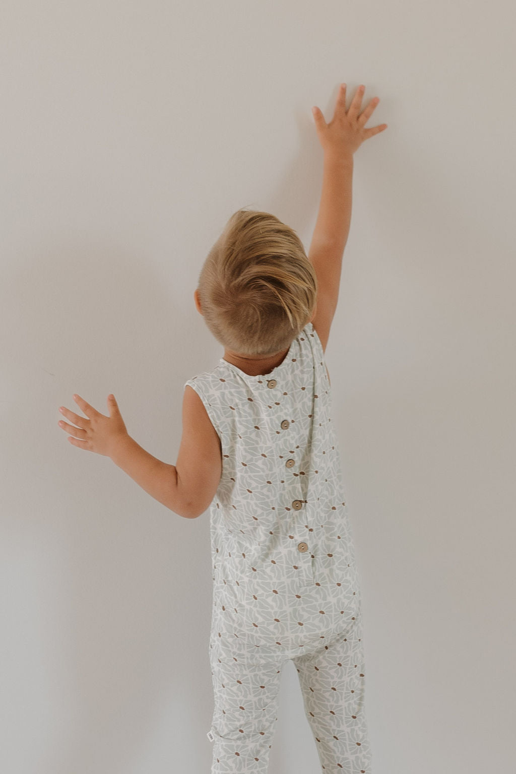 A child with light brown hair is facing a white wall, reaching up with one hand. They are wearing the sleeveless "Harem Bamboo Romper | Coast" by forever french baby, featuring breathable fabric with a patterned design and buttons on the back.