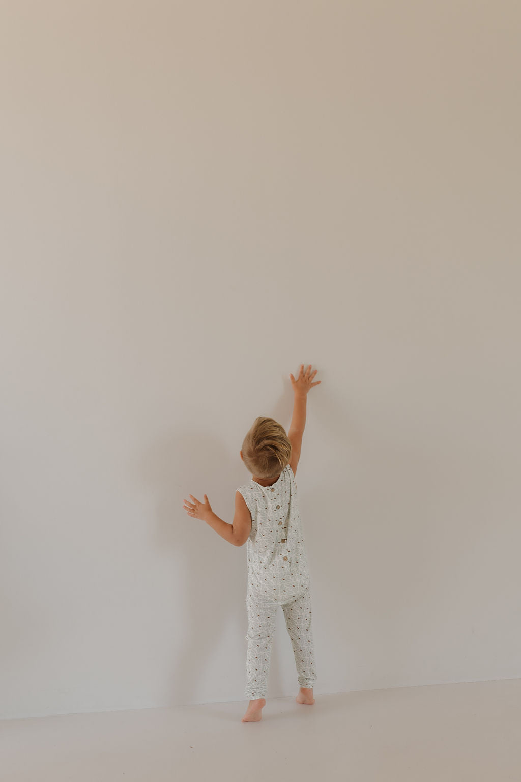 A young child in a "Harem Bamboo Romper | Coast" by forever french baby, crafted from breathable fabric, reaches toward a plain wall. Barefoot on a light-colored floor, the minimalistic scene is softly lit.