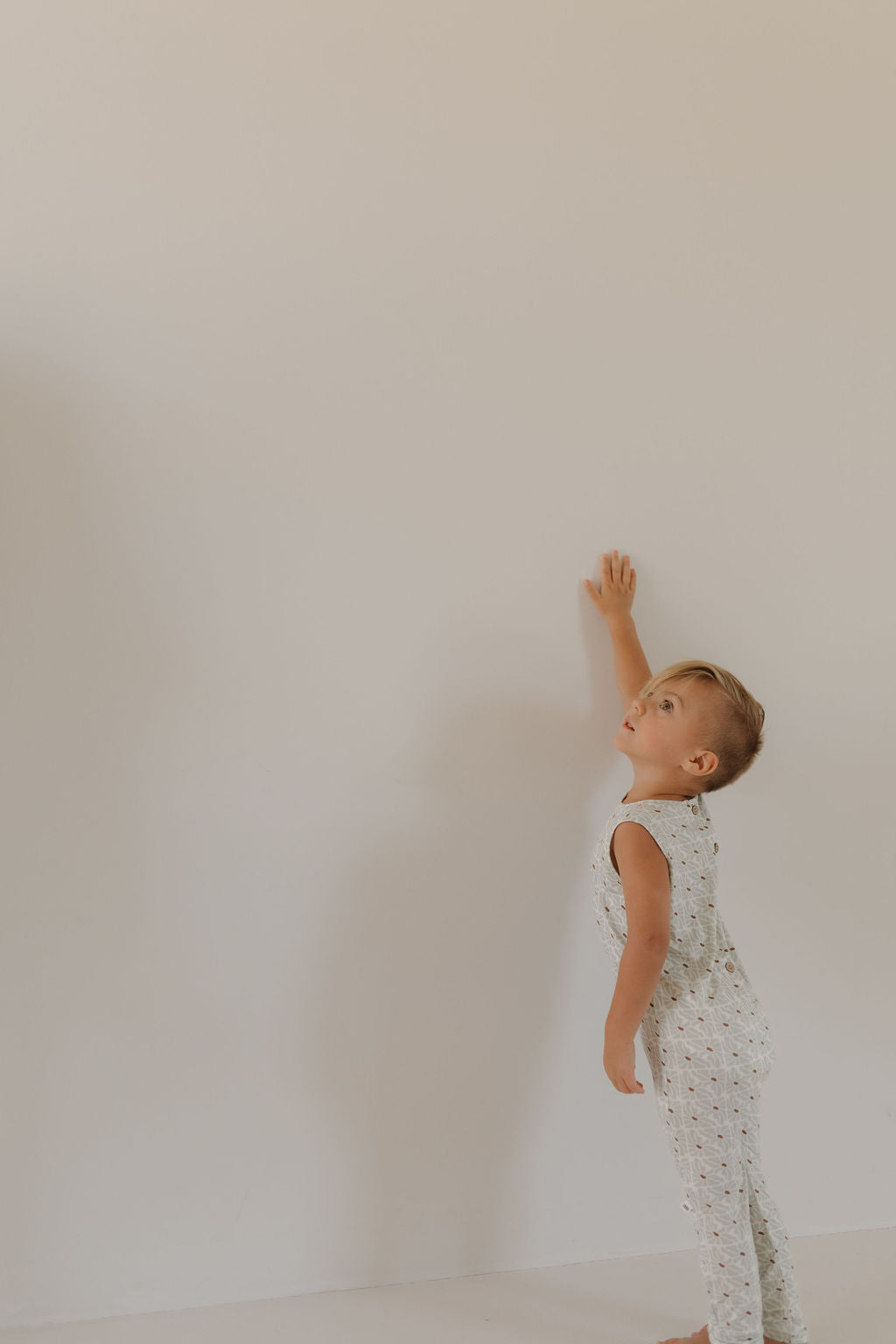 A young child with short hair, in a sleeveless Harem Bamboo Romper by forever french baby, reaches up to a blank white wall indoors, casting a shadow beside them.