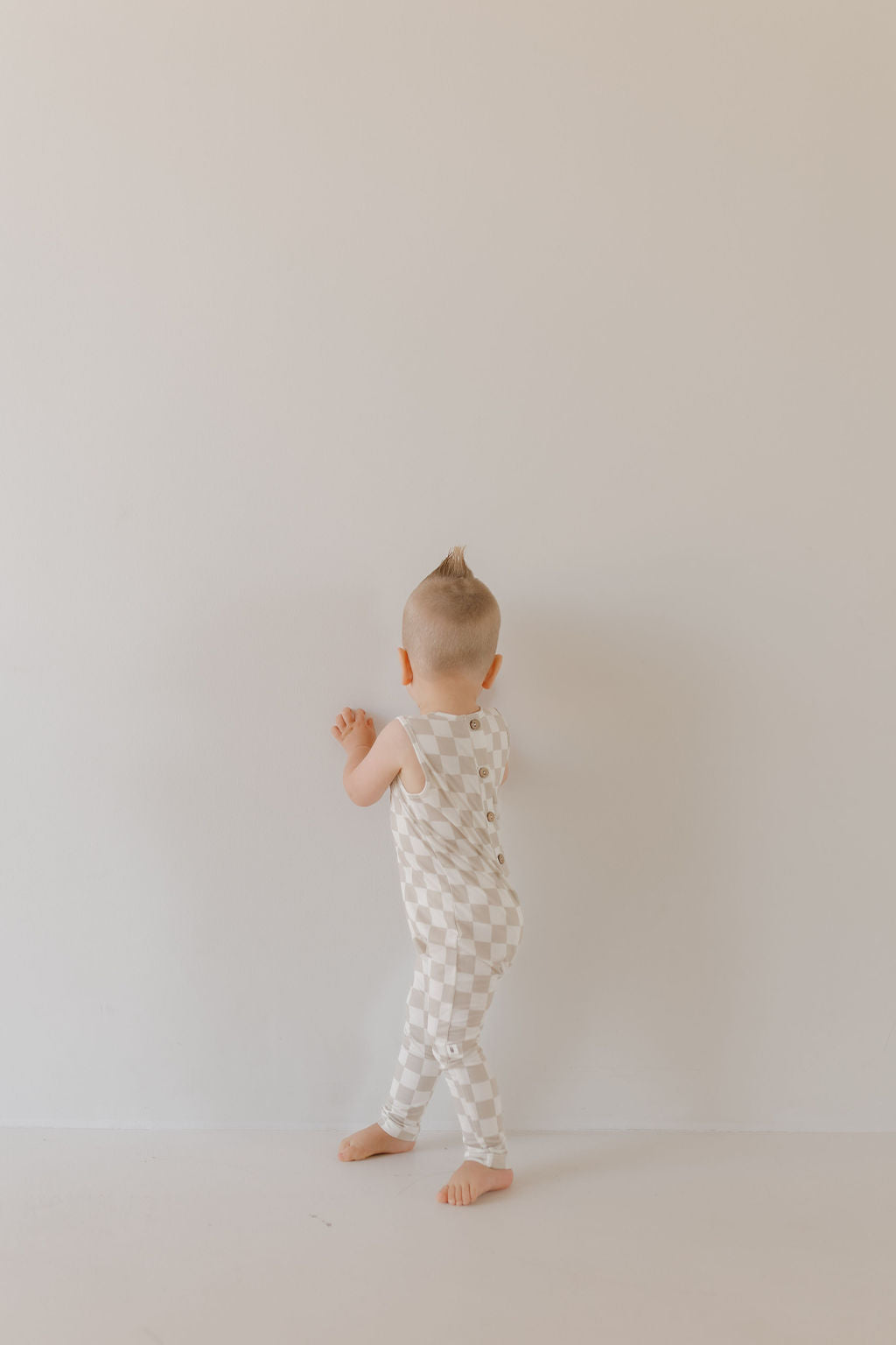 A toddler with a small mohawk wears the Harem Bamboo Romper | Checkerboard by forever french baby, standing barefoot against a plain off-white wall in a minimally decorated room, highlighting the child's playful pose.