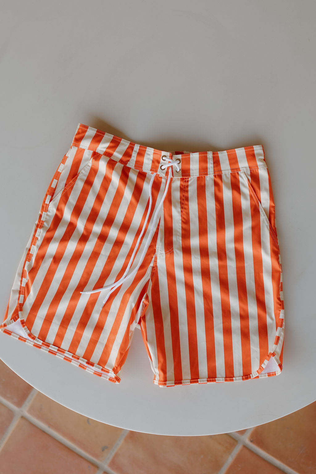 The Men's Boardshort | Poolside by forever french baby, featuring orange and white vertical stripes and a drawstring waist, is laid flat on a round, light-colored table.