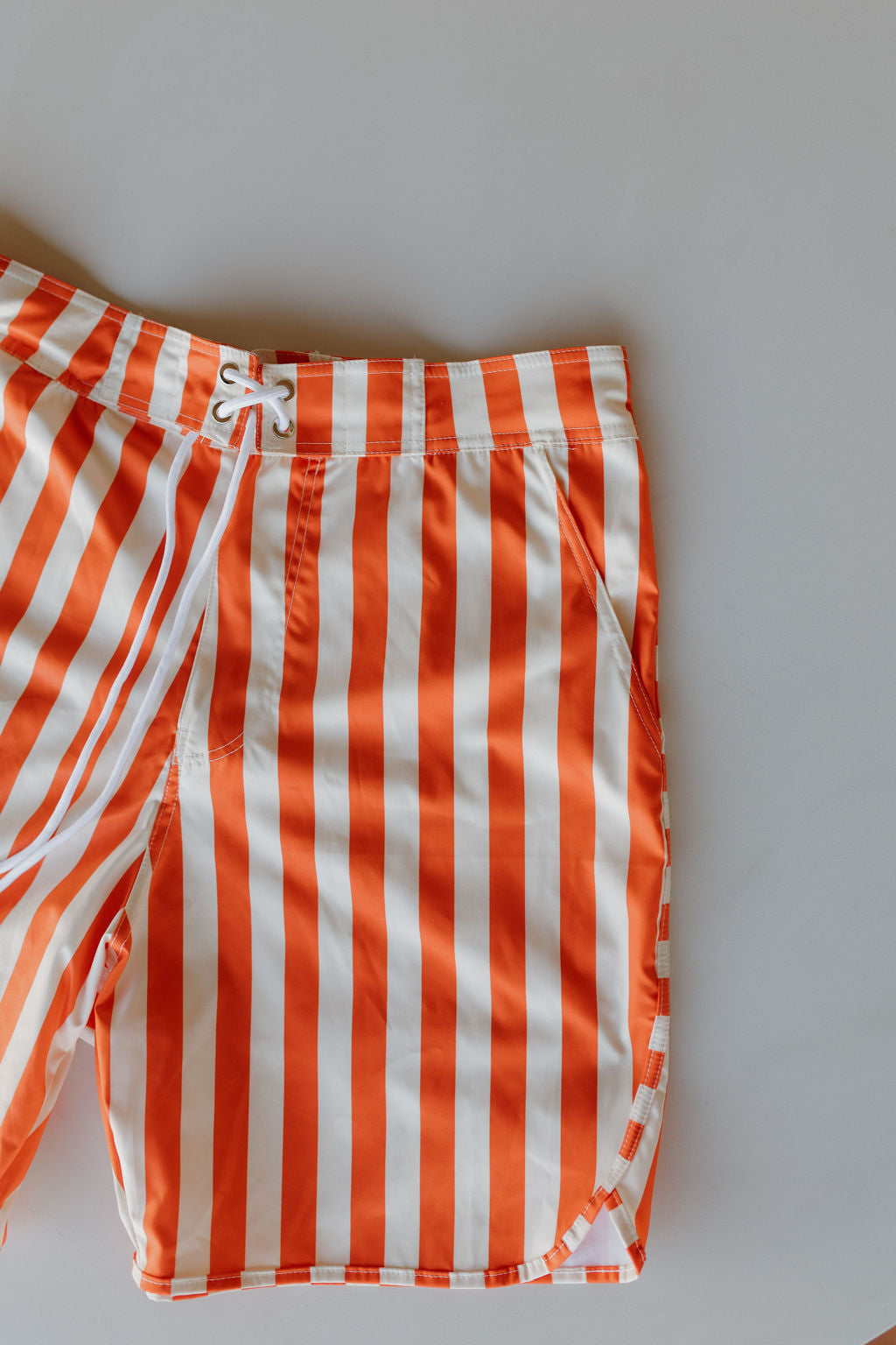 From the Poolside Collection by forever french baby, these men's boardshorts feature orange and white vertical stripes with a white drawstring. The image captures only the right half against a light gray background, emphasizing their contemporary design.
