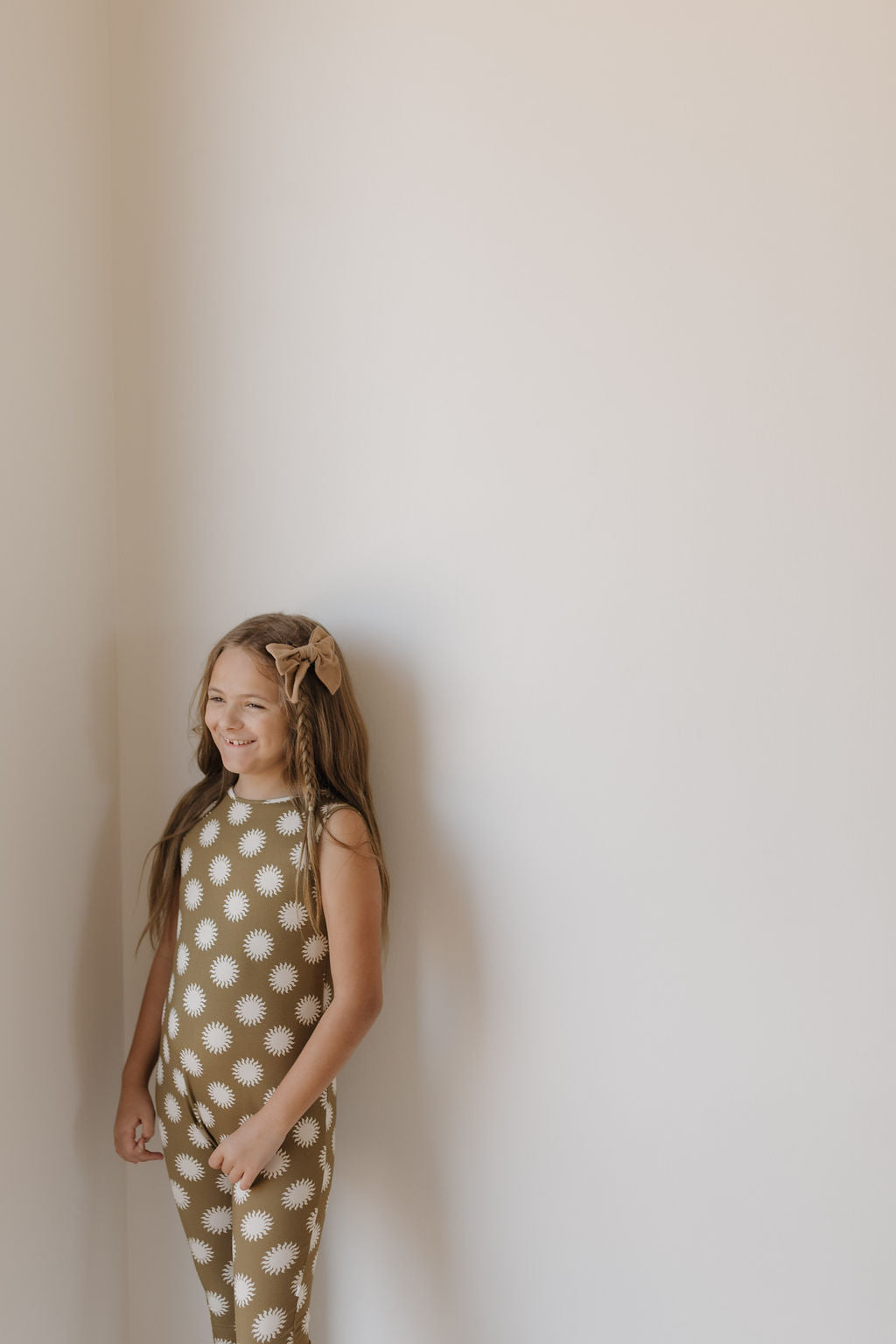 A young girl with long hair stands smiling against a white wall, wearing the Harem Bamboo Romper by forever french baby—a brown outfit adorned with a white floral pattern and featuring a bow in her hair, crafted from breathable bamboo fabric for comfort and style.