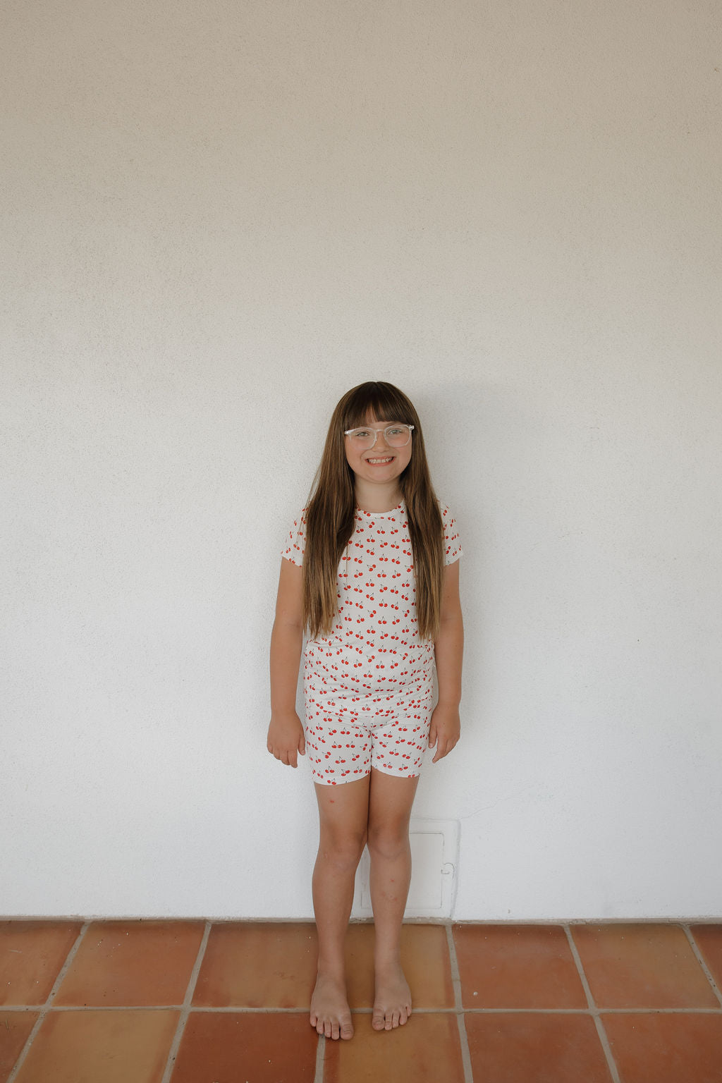 A young girl with long brown hair and glasses stands barefoot on a tiled floor against a white wall, wearing the forever french baby Bamboo Short Set | Sweetheart Cherries—soft, breathable pajamas with a red and blue cherry pattern.