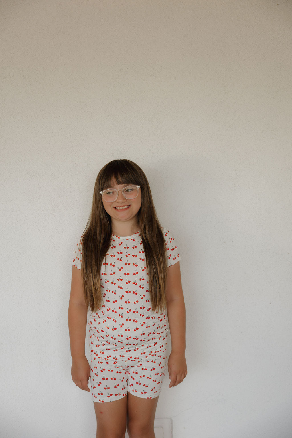 A young girl with long brown hair and glasses smiles against a white wall, wearing the forever french baby Bamboo Short Set | Sweetheart Cherries, and enjoying its super-soft, breathable fabric with a playful red cherry print.