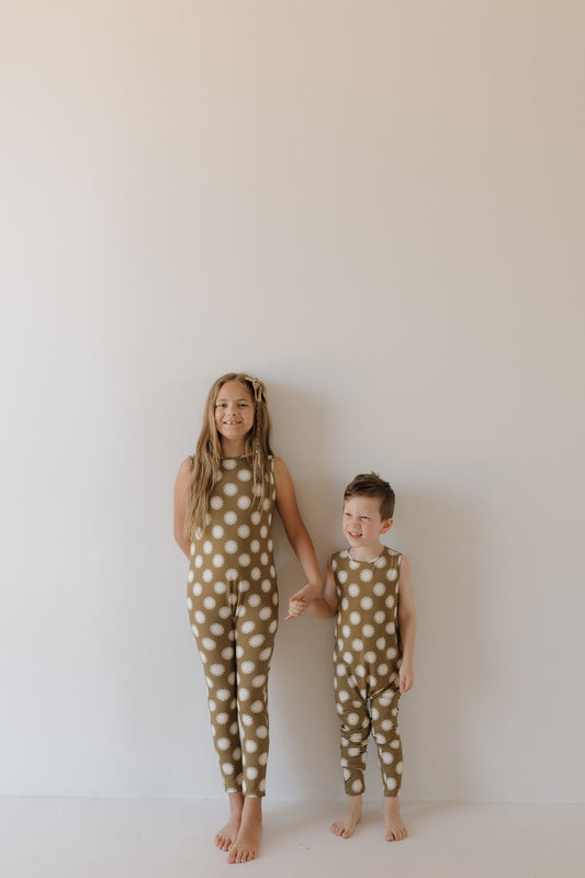 Two children hold hands against a plain backdrop, wearing matching "Harem Bamboo Romper | Sol" outfits by forever french baby. The brown rompers with white polka dots are breathable and hypo-allergenic for comfort. One child has long hair; the other has short locks. Both smile barefoot.