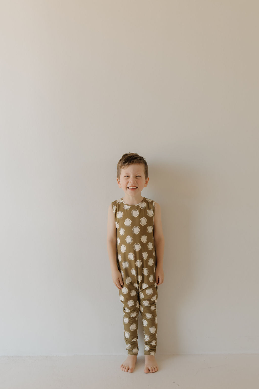 A young child stands barefoot against a plain, light background, smiling in a sleeveless Harem Bamboo Romper | Sol by forever french baby. This olive-green romper with white flowers is hypoallergenic and perfect for sensitive skin, offering an adorable blend of style and comfort.