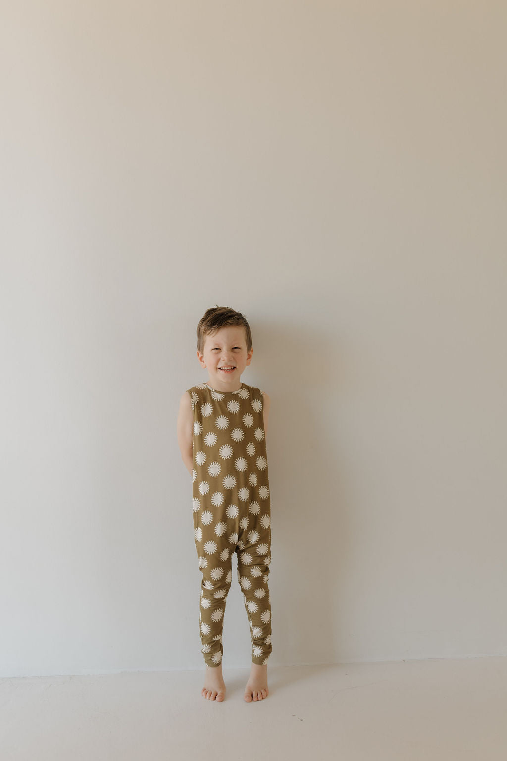 A child stands barefoot against a plain background, wearing the Harem Bamboo Romper | Sol by forever french baby. The green and white polka dot romper is crafted from breathable, hypo-allergenic bamboo for comfort. The child smiles while looking at the camera.