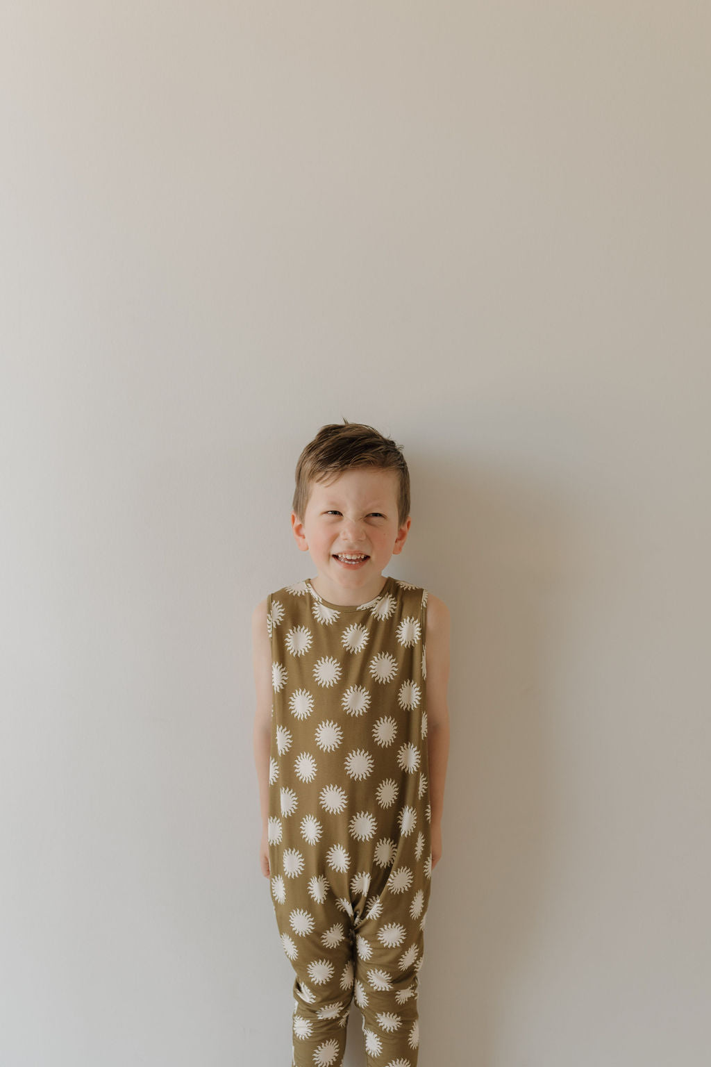 A young child smiles against a plain light background, wearing the sleeveless "Harem Bamboo Romper | Sol" by forever french baby. This stylish romper features white sun/star patterns on brown, crafted from breathable bamboo fabric that covers the child from shoulders to ankles.
