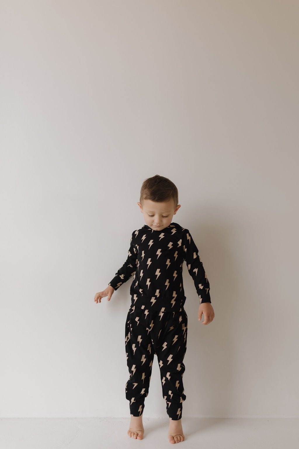 A young child stands against a plain, light background wearing the "Hooded Set | Midnight Lightning Bolt" by forever french baby, featuring a black cotton-spandex onesie with a lightning bolt print. The child's arms are slightly raised as they look down.