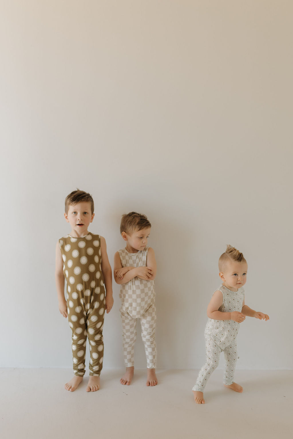Against a plain backdrop, three young children stand barefoot. The first wears the Harem Bamboo Romper in green by "forever french baby," crafted from breathable bamboo fabric with white patterns. The second sports a beige checkered outfit with folded arms, and the third dons a white ensemble with small designs.