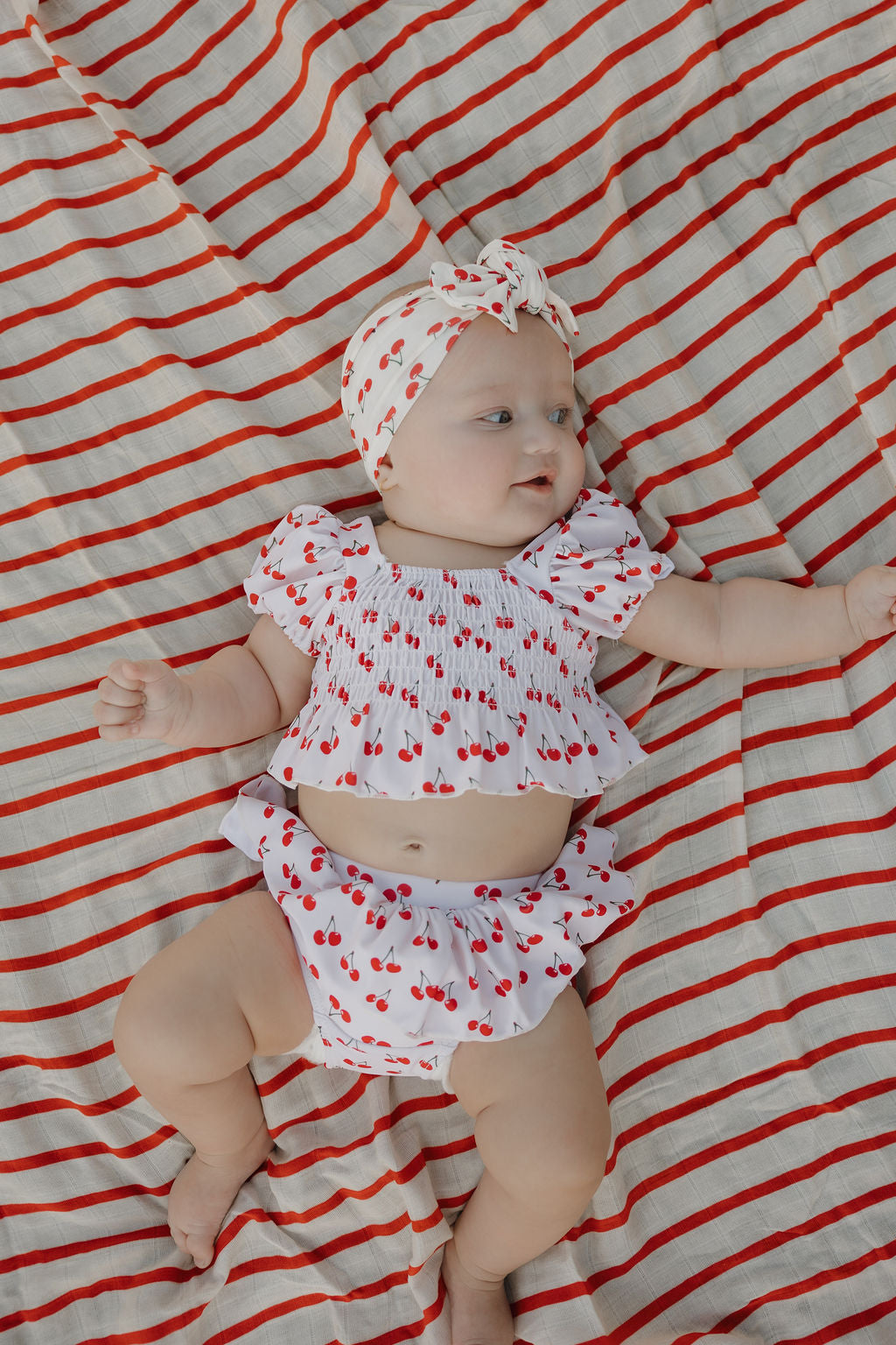 A baby wears the forever french baby Toddler Two Piece Swimsuit | Sweetheart Cherries with a matching headband, lying on a red-and-white striped blanket and looking to the side.
