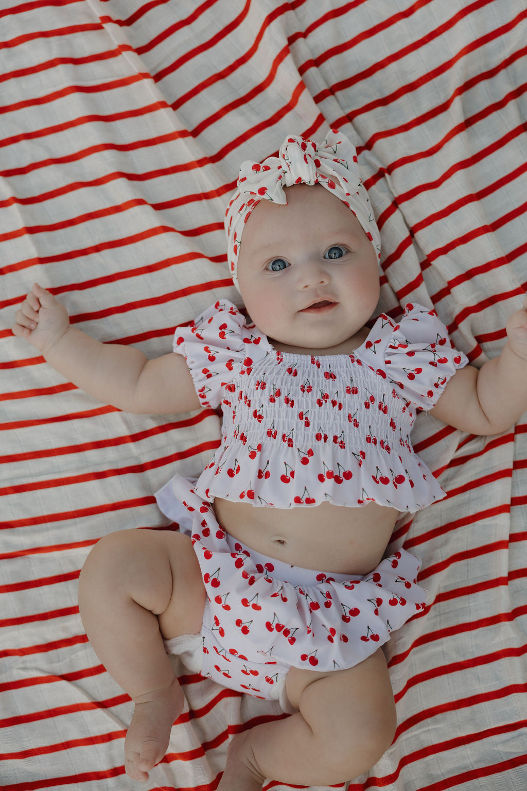 A blue-eyed baby lies on a red-and-white striped blanket, wearing a cherry-print ruffled outfit and the Forever French Baby Bamboo Head Wrap in Sweetheart Cherries, with arms slightly raised and gazing up.