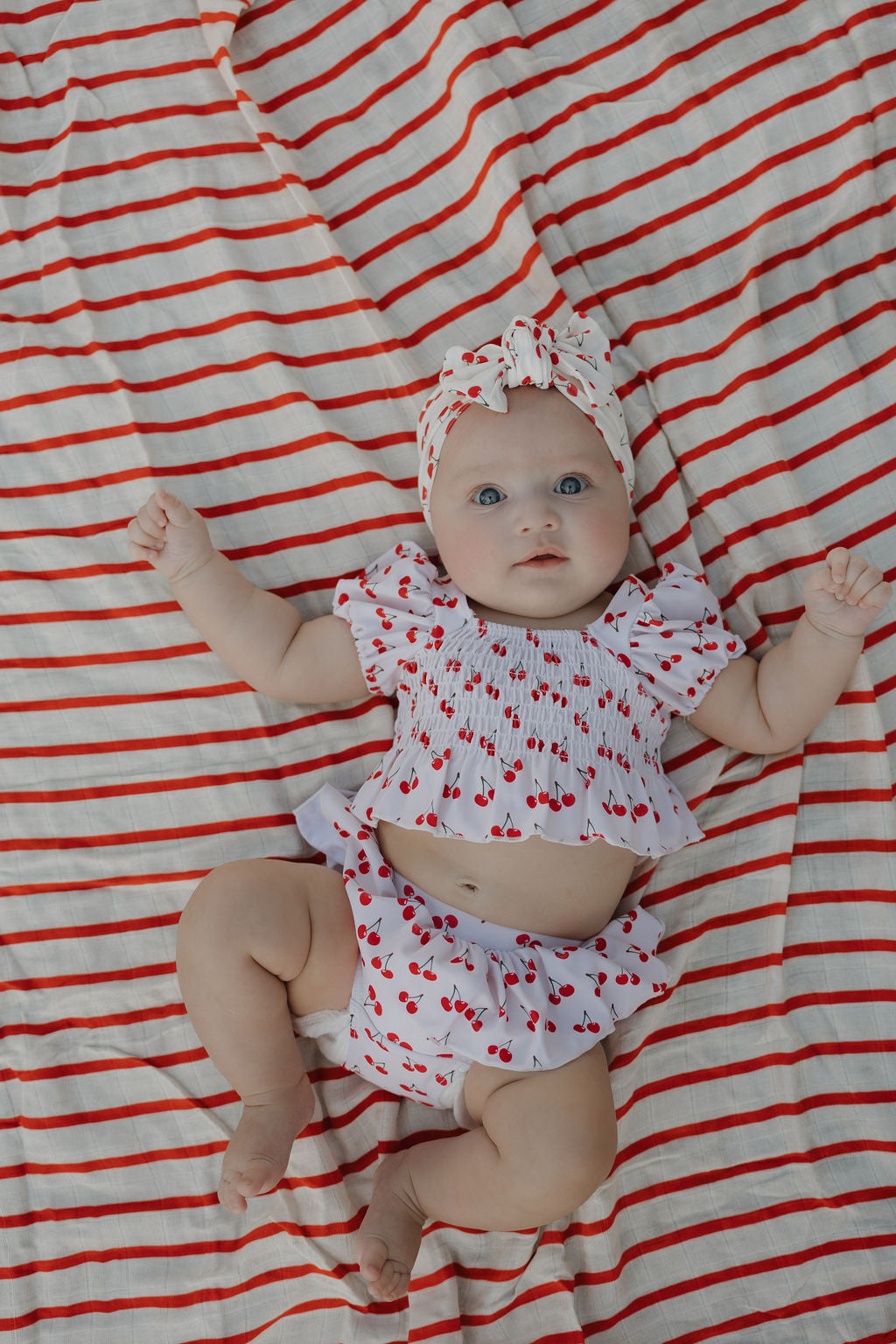 A baby in a Sweetheart Cherries bamboo head wrap from forever french baby lies on a white and red-striped blanket, looking up with arms raised—a sweet moment.