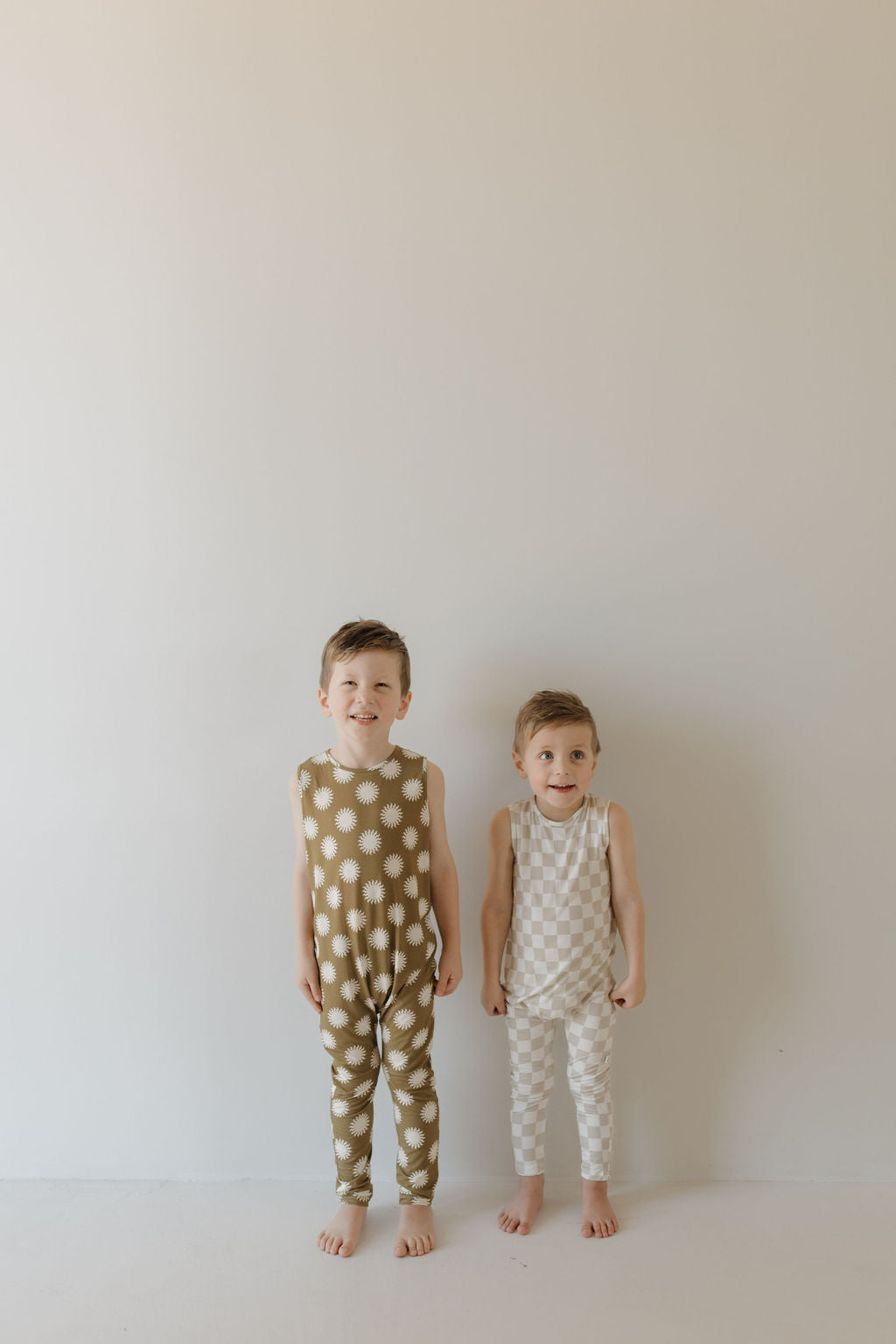 Two young children stand barefoot against a plain background, both smiling in sleeveless polka dot Harem Bamboo Rompers by forever french baby—one in green and white, the other in beige and white. Crafted from breathable bamboo fabric, these rompers keep them comfy all day long.