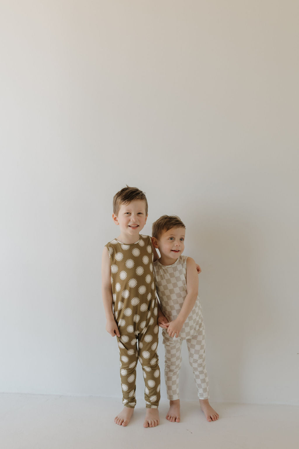 Two kids stand barefoot by a white wall; one in green with white stars, and the other in a beige checkered design. Both smile while wearing hypoallergenic bamboo outfits by forever french baby, including the Harem Bamboo Romper | Sol.