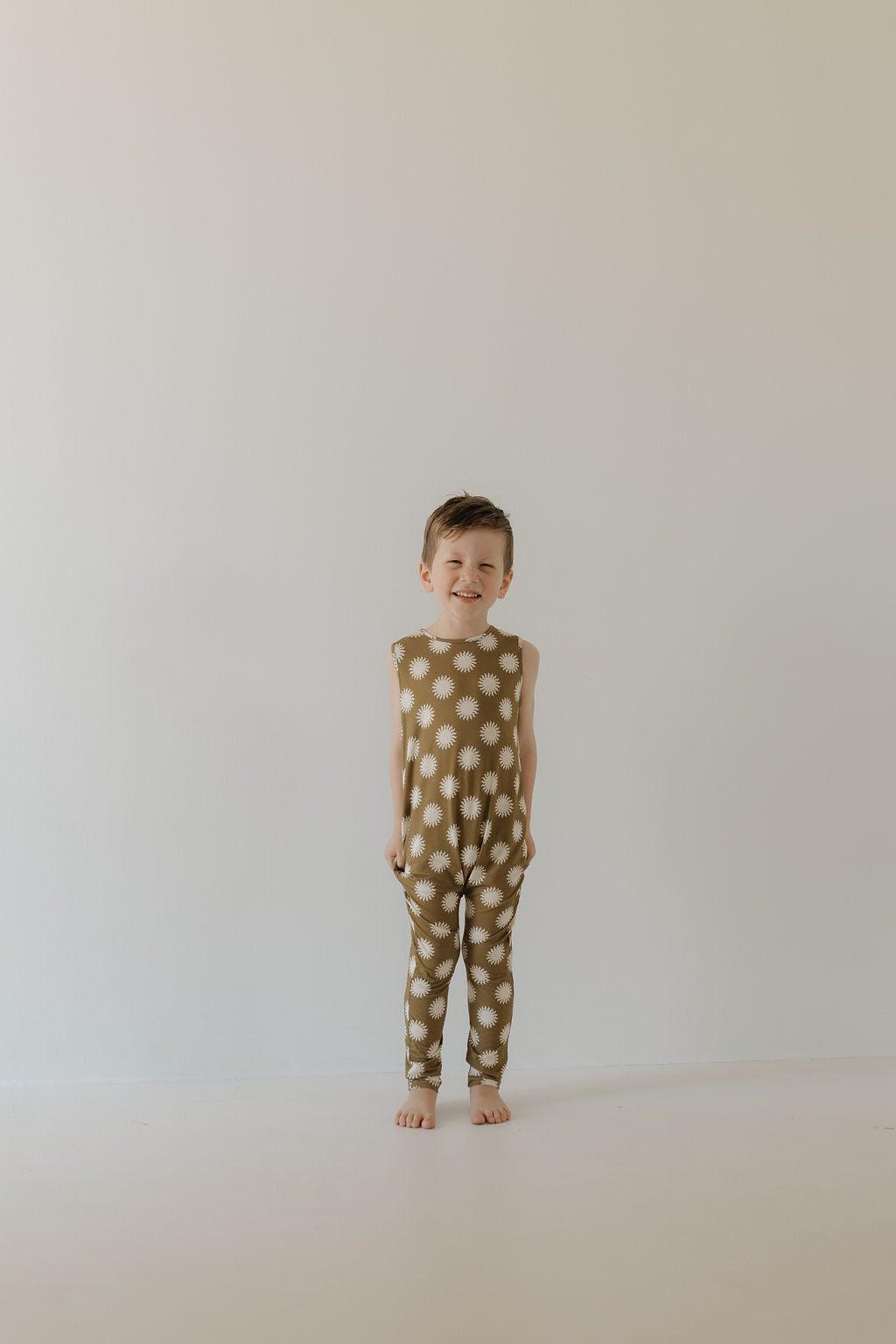 A child beams barefoot against a plain backdrop, wearing the forever french baby Harem Bamboo Romper | Sol. The sleeveless romper features a white starburst design on an olive background and is made from breathable bamboo fabric for comfort and style.