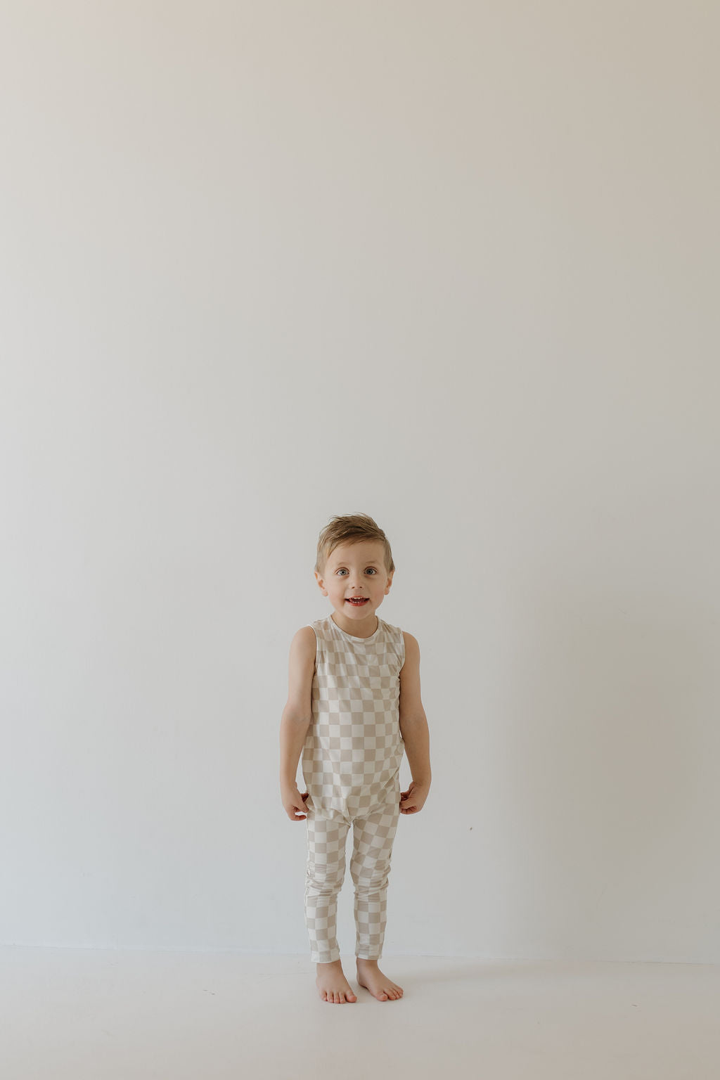 A young child stands barefoot against a plain background, wearing the sleeveless Harem Bamboo Romper | Checkerboard from forever french baby. The child looks directly at the camera with a neutral expression, hands relaxed by their sides.