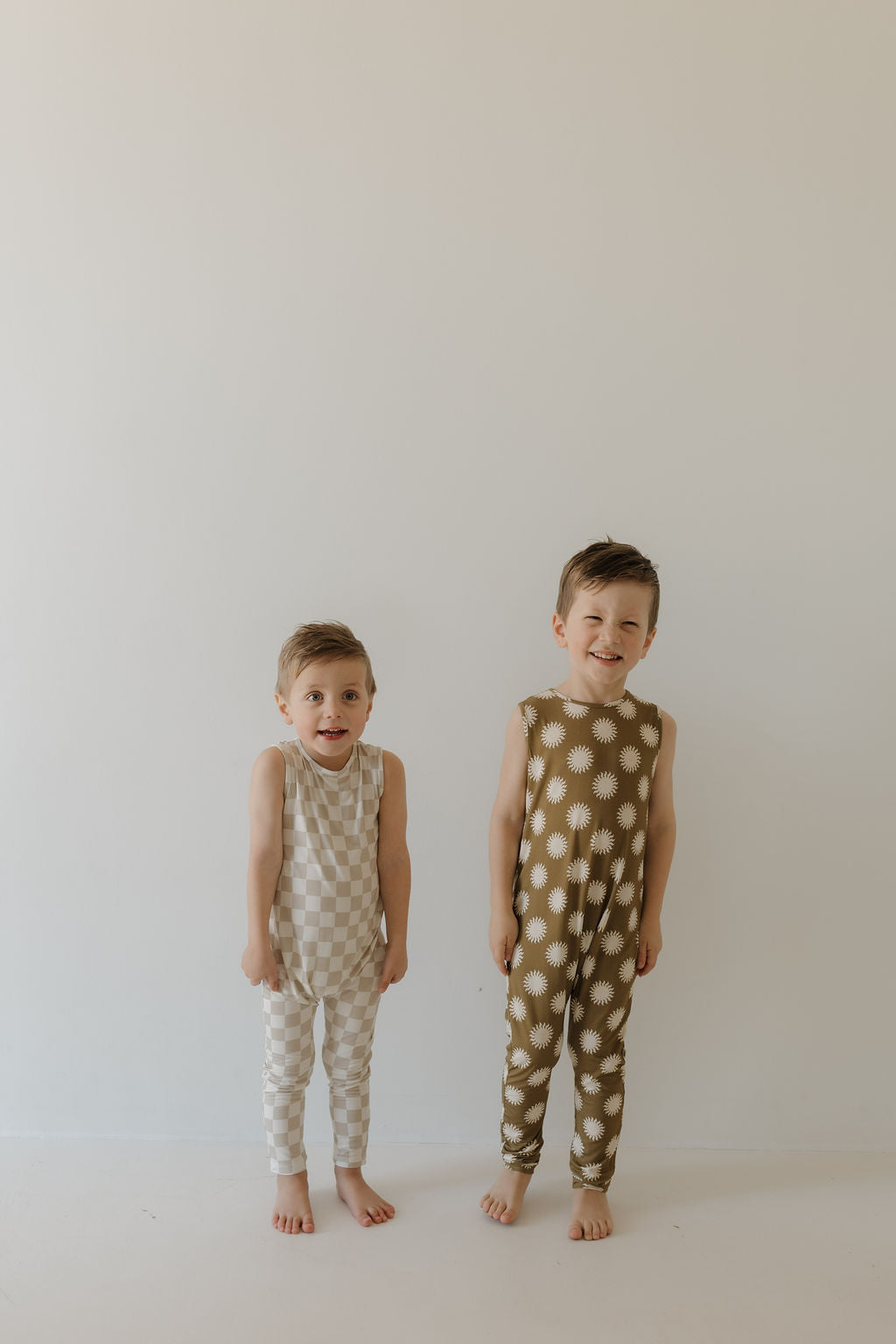 Two children stand barefoot on a white background. One wears a light checkered outfit, while the other dons the Harem Bamboo Romper | Sol by forever french baby with white sun patterns. Made from breathable bamboo fabric, both outfits enhance their short hair and bright smiles.