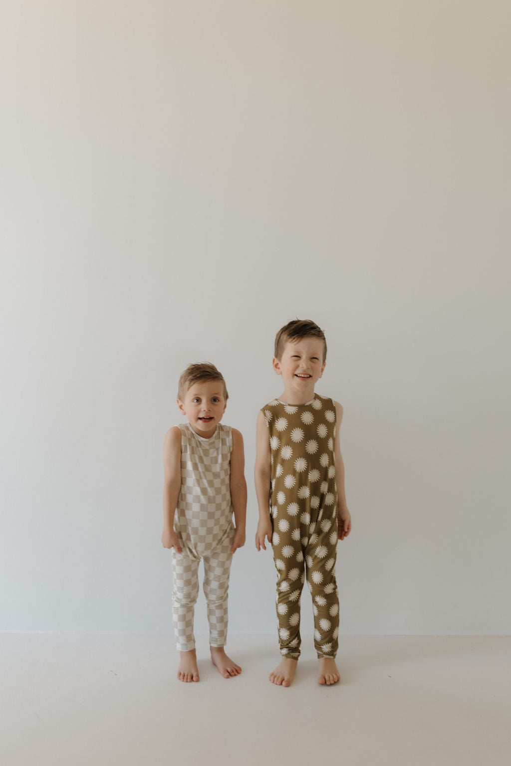 Two young children stand barefoot against a plain background in sleeveless harem bamboo rompers from forever french baby, one beige with polka dots and the other brown. They face the camera, with one child smiling broadly.