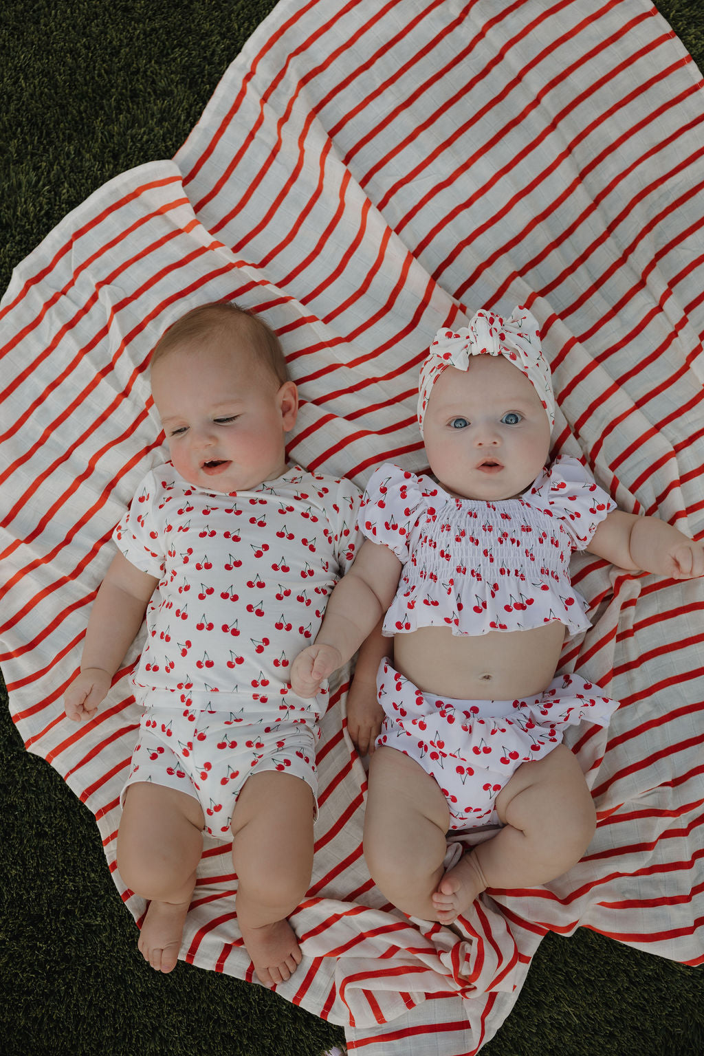 Two babies relax side by side outdoors on a red-and-white striped blanket, both wearing forever french baby Toddler Two Piece Swimsuit | Sweetheart Cherries; the baby on the right also wears a matching headband.