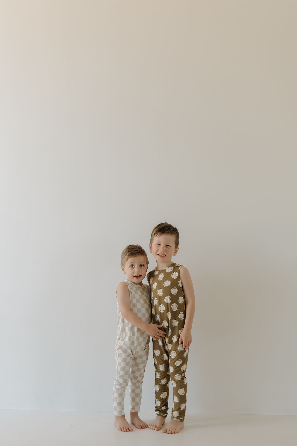 Two happy barefoot children stand in a minimal setting. The left child wears a light checkered outfit, while the right one is dressed in the "Harem Bamboo Romper | Sol" by forever french baby, featuring a dark star pattern and crafted from breathable bamboo fabric against a light background.