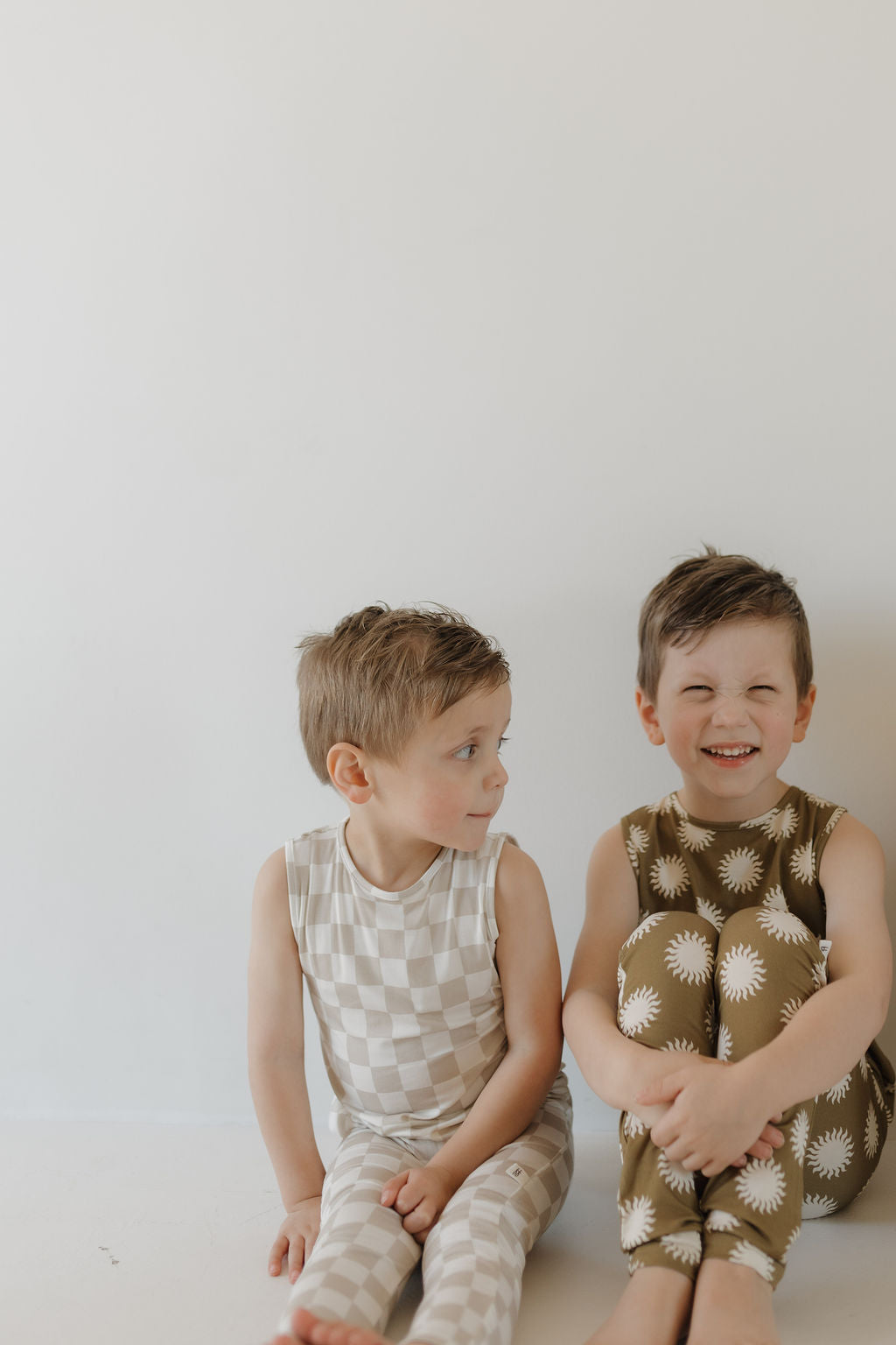 Two young children sit comfortably on a light background. One wears a beige checkered outfit, while the other beams at the camera in forever french baby's Harem Bamboo Romper | Sol, featuring white sun patterns for stylish and hypo-allergenic comfort.