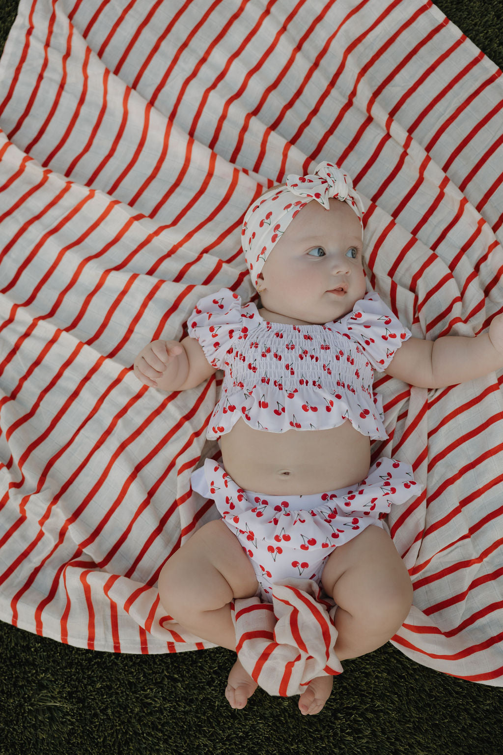 A baby in a Forever French Baby Bamboo Head Wrap | Sweetheart Cherries lies on a red-and-white striped blanket on the grass, gazing to the side with one arm raised.