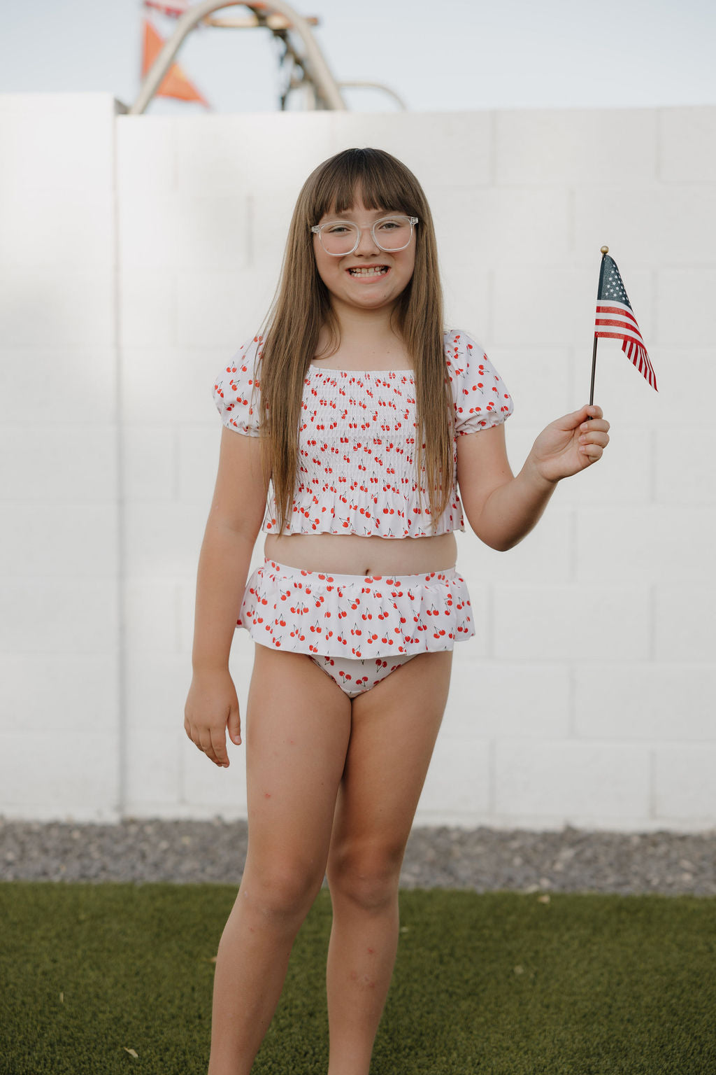 A young girl with long brown hair and glasses stands on grass, smiling and holding a small American flag. She wears the forever french baby Toddler Two Piece Swimsuit | Sweetheart Cherries, featuring a cute cherry print.
