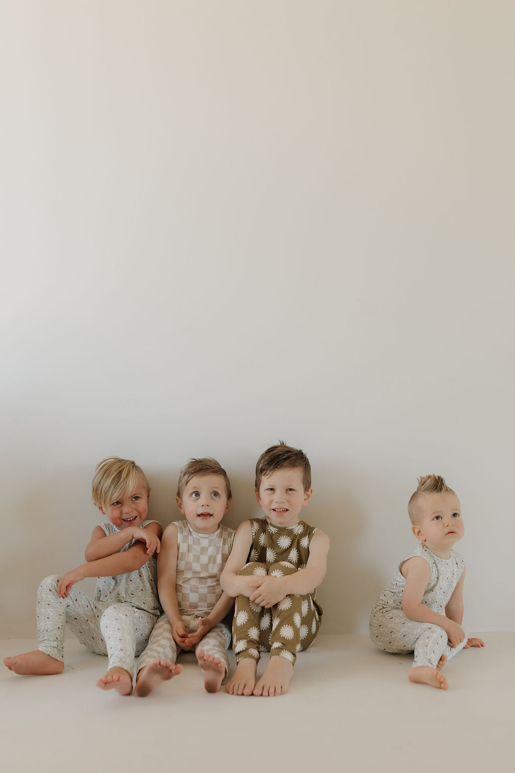 Four toddlers sit on the floor against a plain wall, each wearing the Harem Bamboo Romper | Sol from forever french baby. These breathable rompers feature unique designs. Their expressions vary from curious to playful, capturing childhood wonder perfectly.