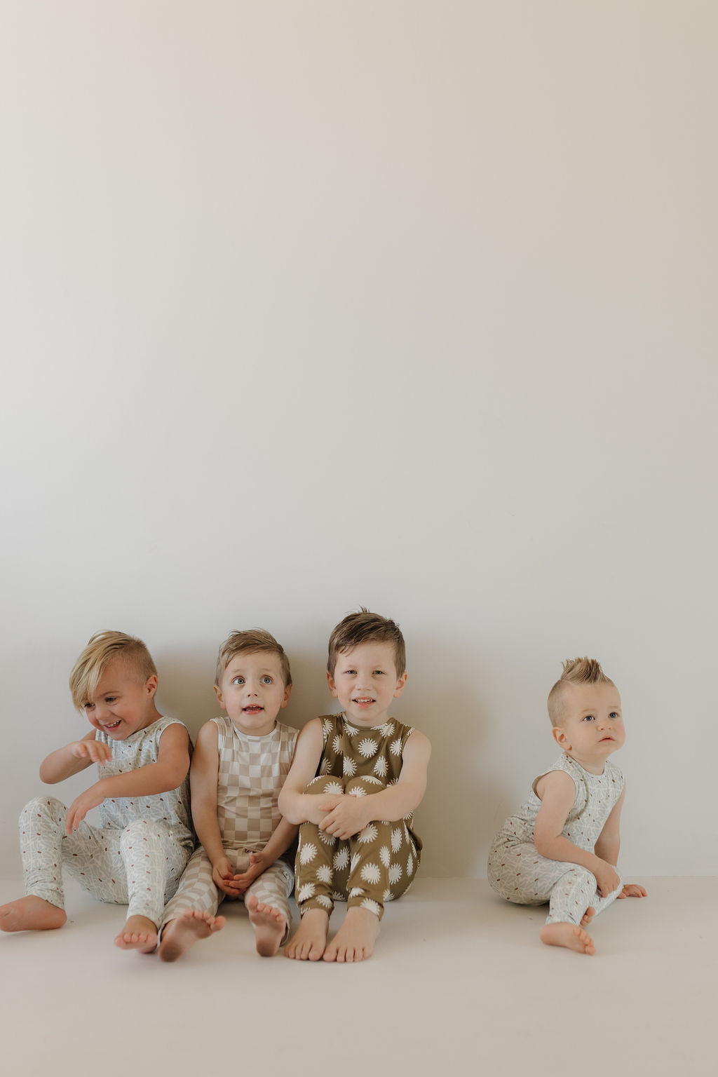 Four smiling young children, barefoot with varied hairstyles, sit against a simple wall in forever french baby's hypo-allergenic Harem Bamboo Romper | Sol jumpsuits. The breathable bamboo fabric ensures comfort and style.