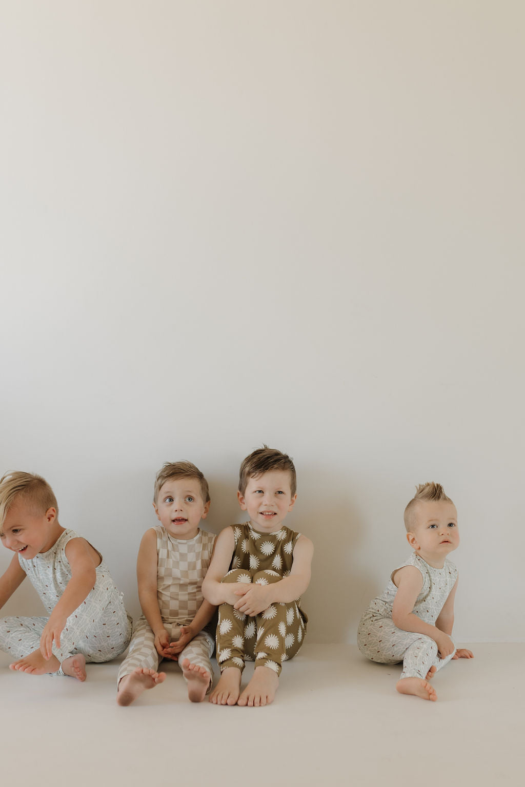 Four young children sit against a plain background, each wearing a unique Harem Bamboo Romper | Sol by forever french baby. Their short hair and barefoot poses highlight their expressive faces while the breathable bamboo fabric ensures comfort.