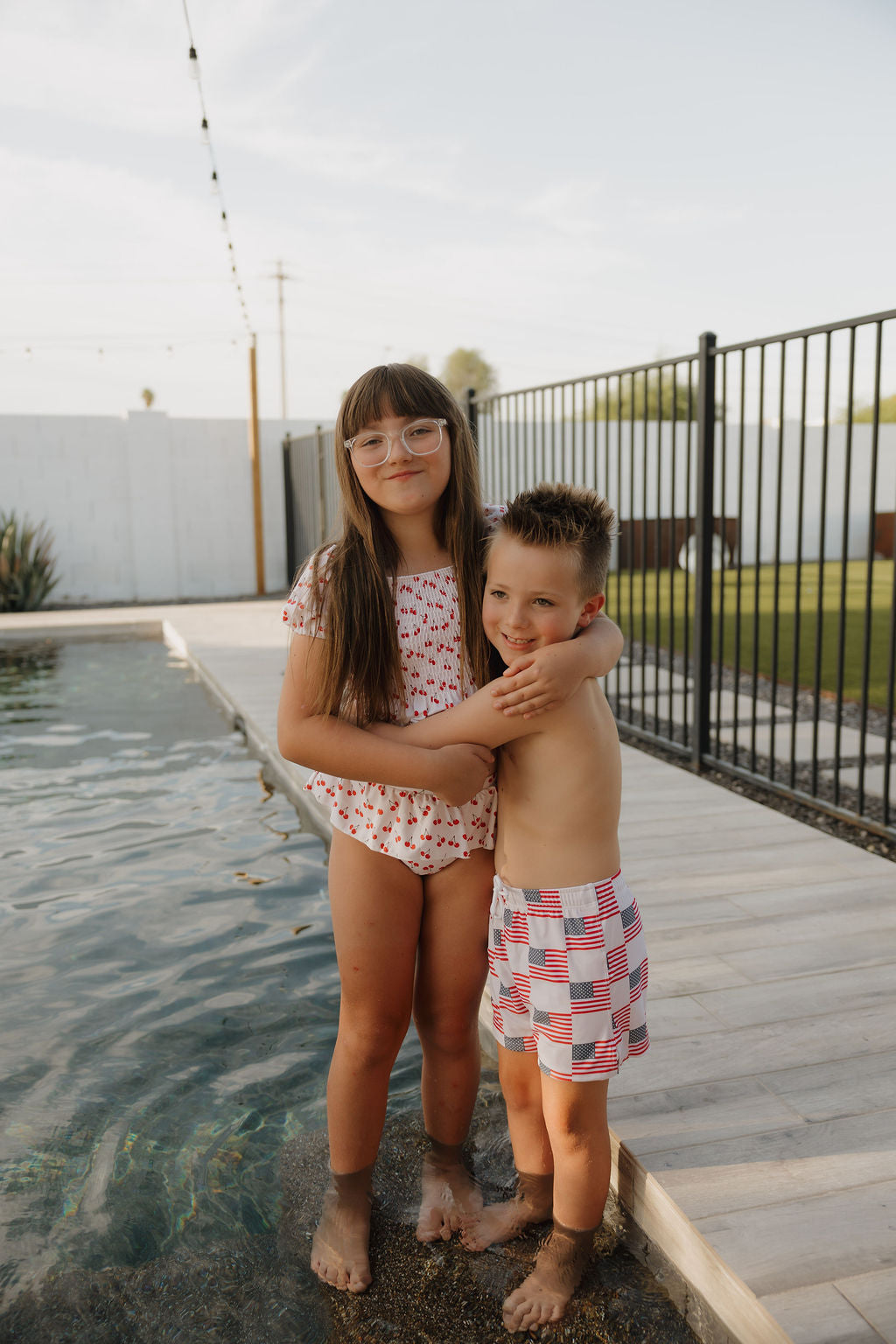 Two young children smile by a pool; the girl, in forever french baby's Toddler Two Piece Swimsuit | Sweetheart Cherries, hugs a boy in flag trunks. Both are outdoors, with grass and a fence behind them.