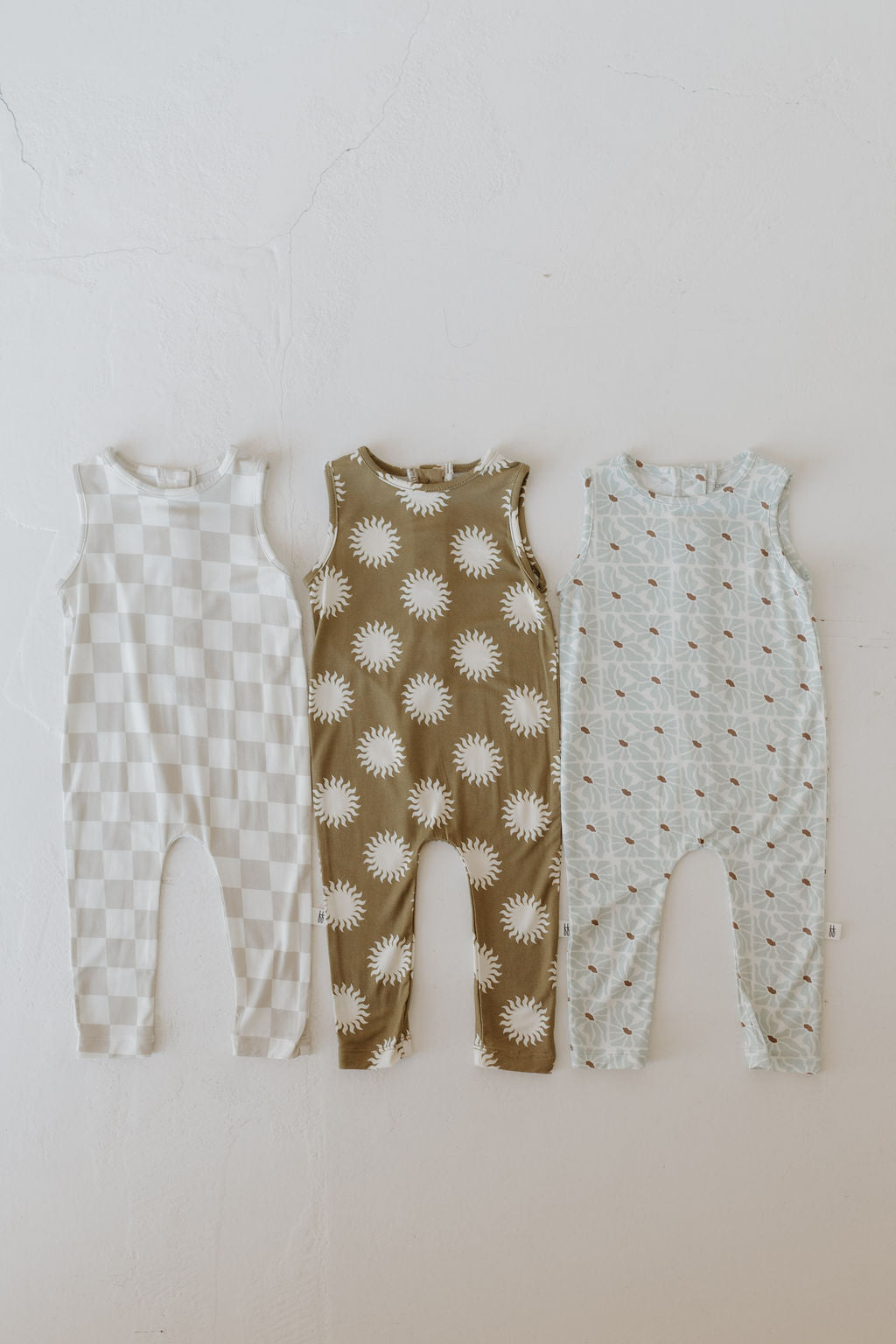 The display shows three baby rompers: the left is beige and white checkered, crafted from breathable fabric; the middle is olive with white suns, and the right is light blue with a leaf design. The center romper is the Harem Bamboo Romper | Sol by forever french baby.