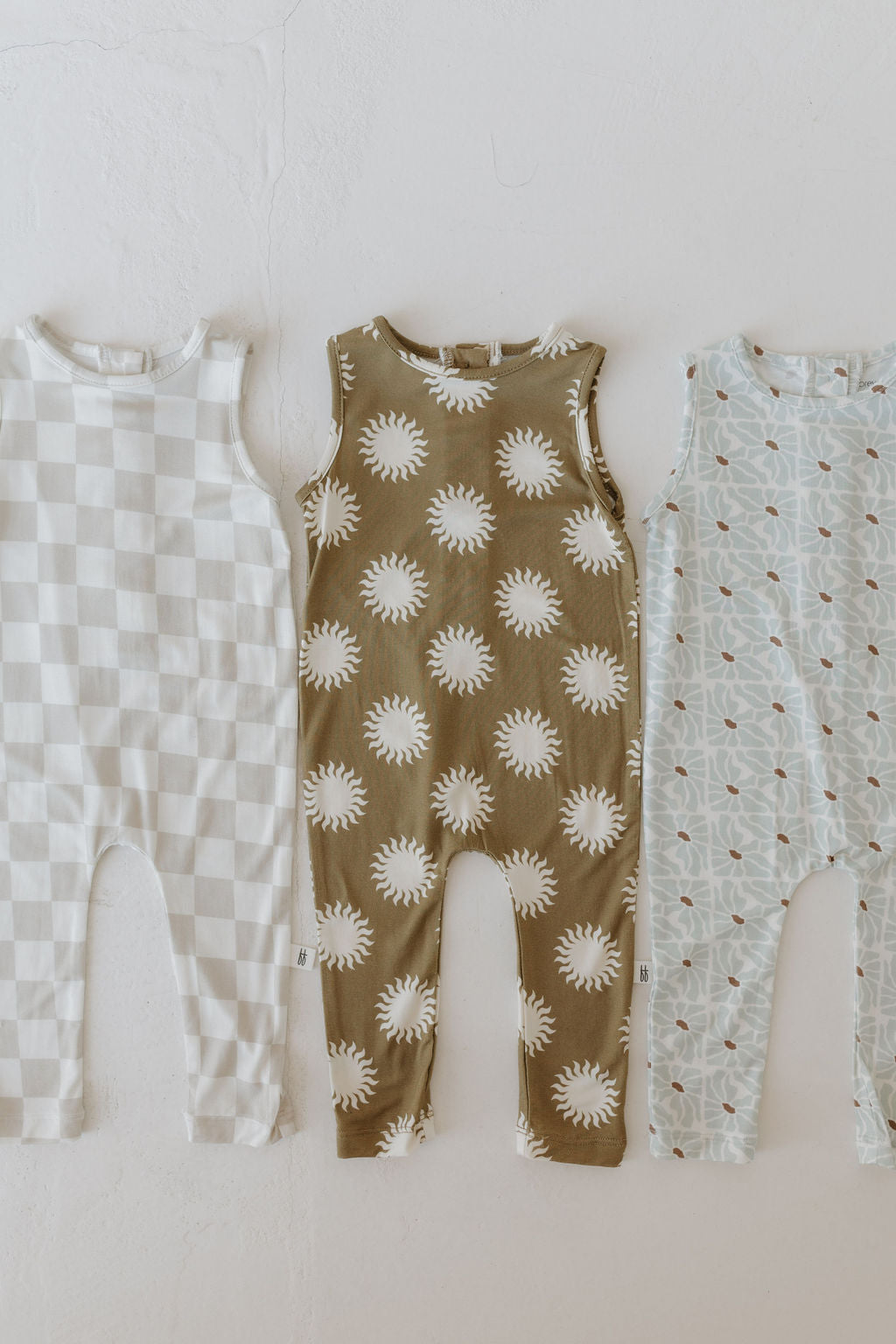 Three baby rompers from forever french baby are displayed against a white wall. The Harem Bamboo Romper | Sol features three designs: checkered, sun pattern, and abstract shapes. Crafted from breathable bamboo, each romper is sleeveless and hypo-allergenic for sensitive skin comfort.