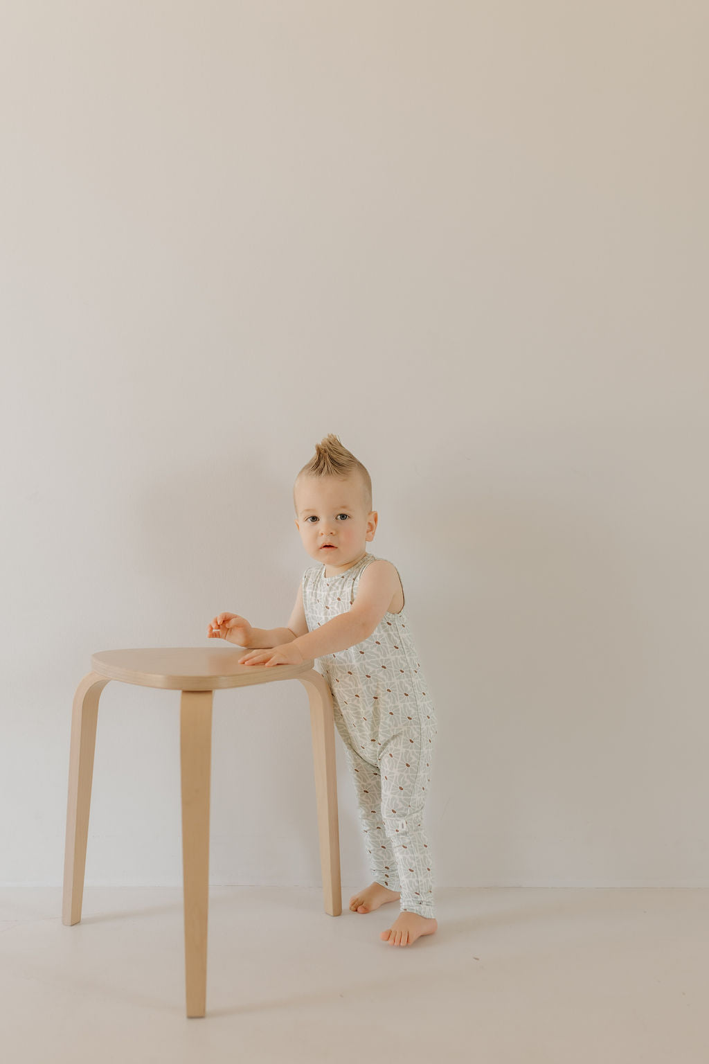 A toddler with a small mohawk stands by a wooden stool, wearing the sleeveless Harem Bamboo Romper in Coast by forever french baby. They gaze toward the camera against a plain, light wall. The romper's breathable bamboo fabric ensures comfort and is perfect for sensitive skin.