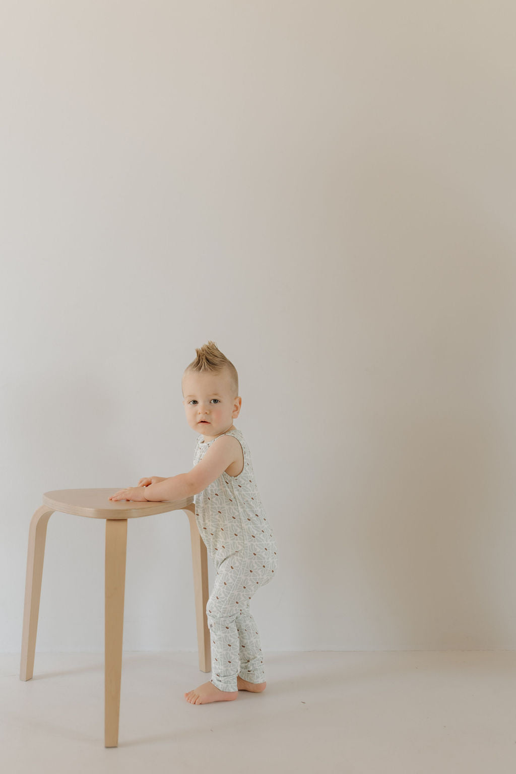 A toddler wearing a "Harem Bamboo Romper | Coast" by forever french baby stands next to a small, light wooden table. The child sports a fauxhawk and looks at the camera against a plain, neutral wall that complements the hypo-allergenic, breathable fabric of the romper.