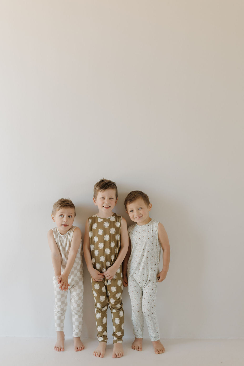 Three young children stand against a plain wall, smiling in their matching forever french baby Harem Bamboo Rompers | Sol. The breathable outfits feature charming patterns: white with gray dots, brown with white stars, and white adorned with tiny black motifs.