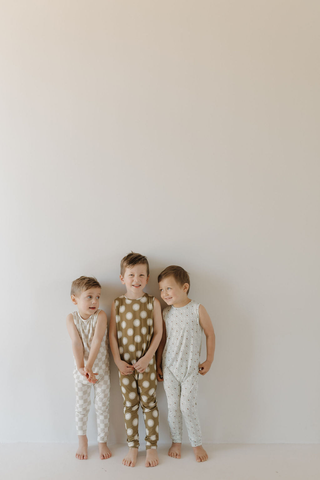 Three young children stand smiling against a plain wall in their Harem Bamboo Romper | Sol by forever french baby. These sleeveless rompers, adorned with polka dots and other patterns, are crafted from breathable bamboo fabric for all-day comfort.