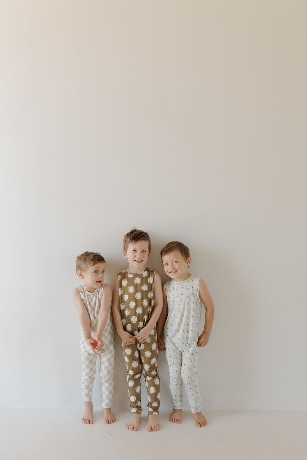 Three young children stand against a plain wall in breathable Harem Bamboo Rompers by forever french baby. The playful outfits feature patterns like brown with large polka dots, white with small dots, and white with checkered squares. Hypo-allergenic and barefoot, they smile cheerfully.